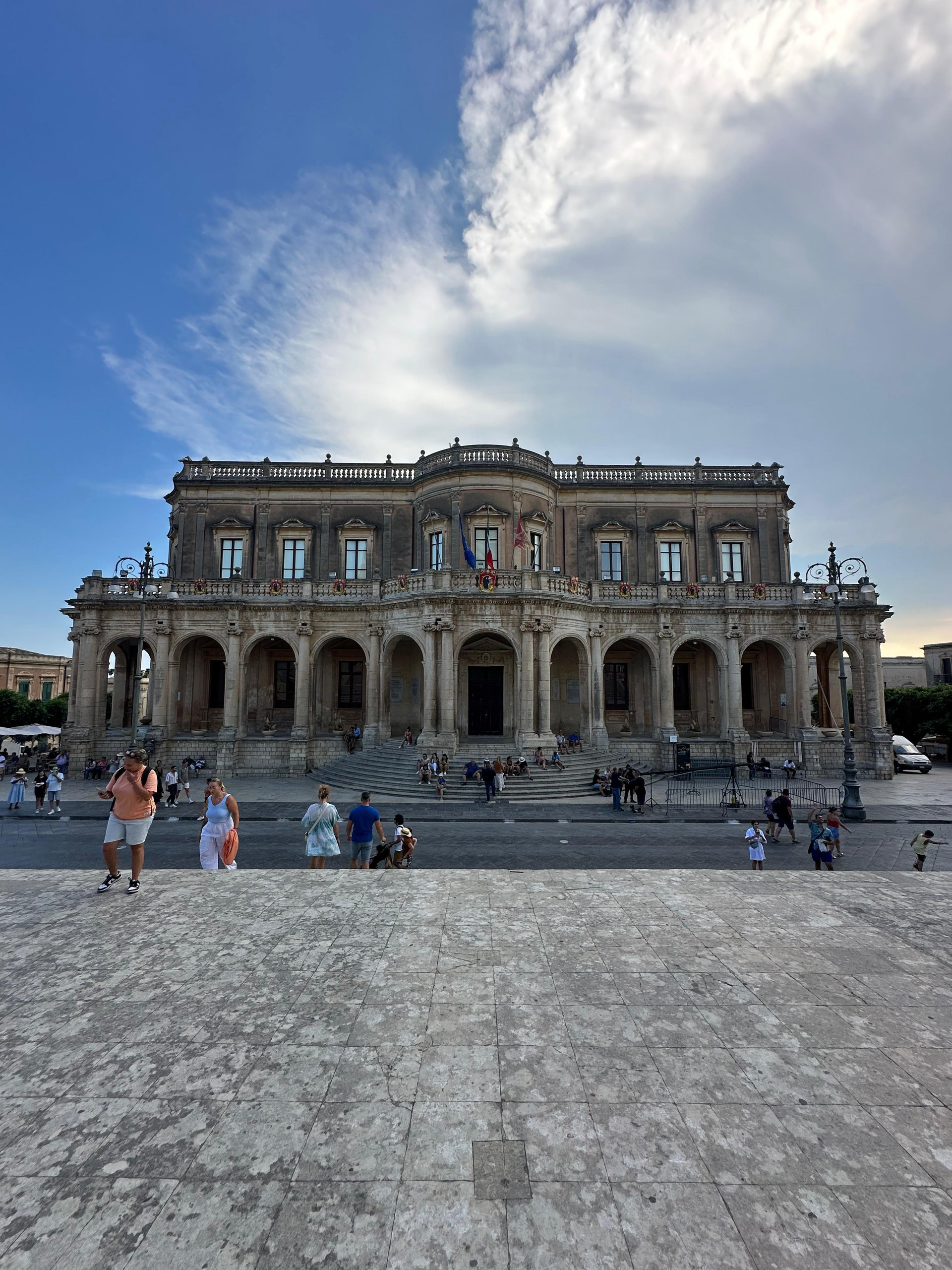 Baroque palace with a curved balustraded facade on a sunlit square under a dramatic sky in Noto