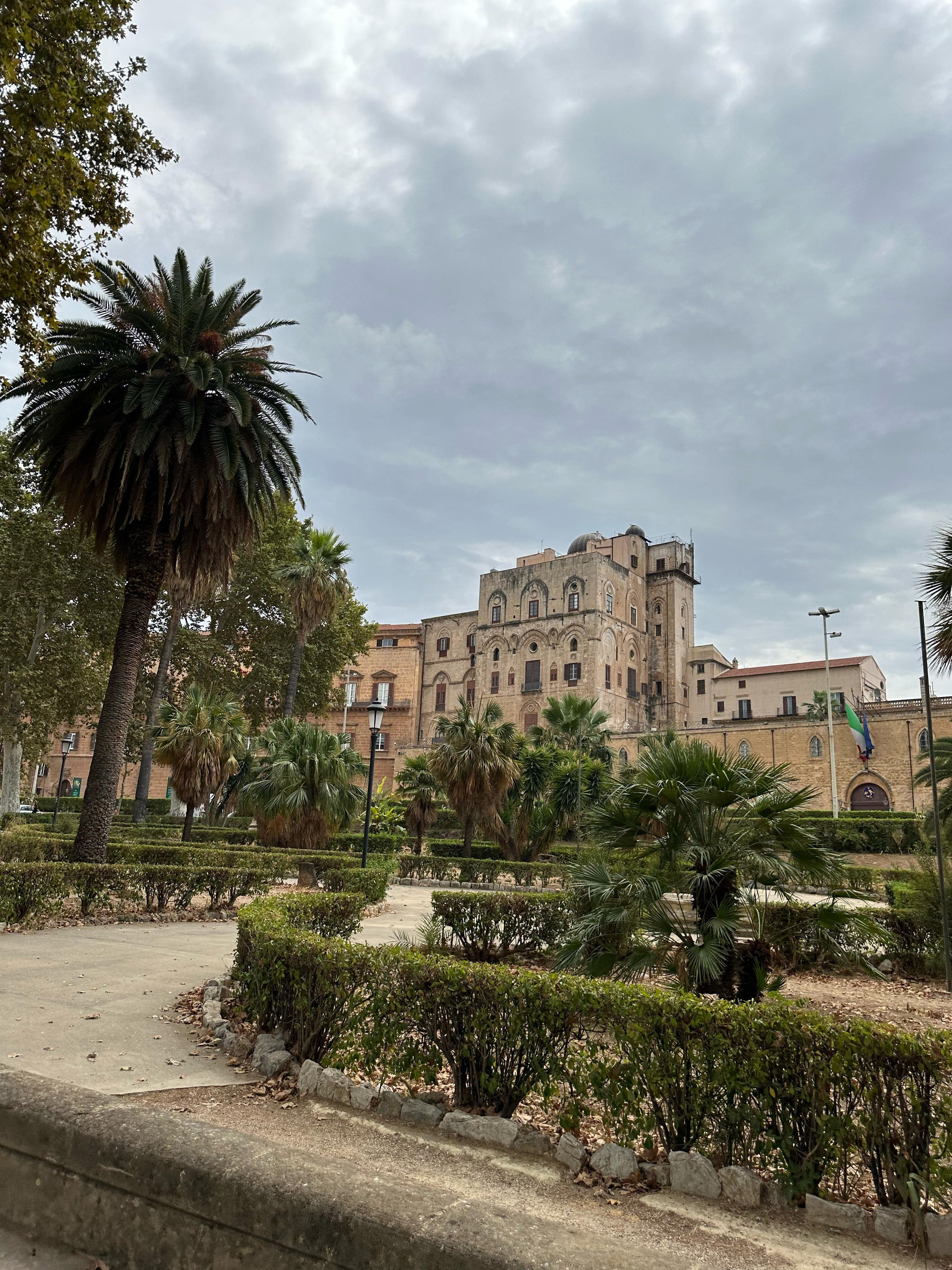 Norman palace towering behind palm trees and clipped hedges under a cloudy sky in Palermo