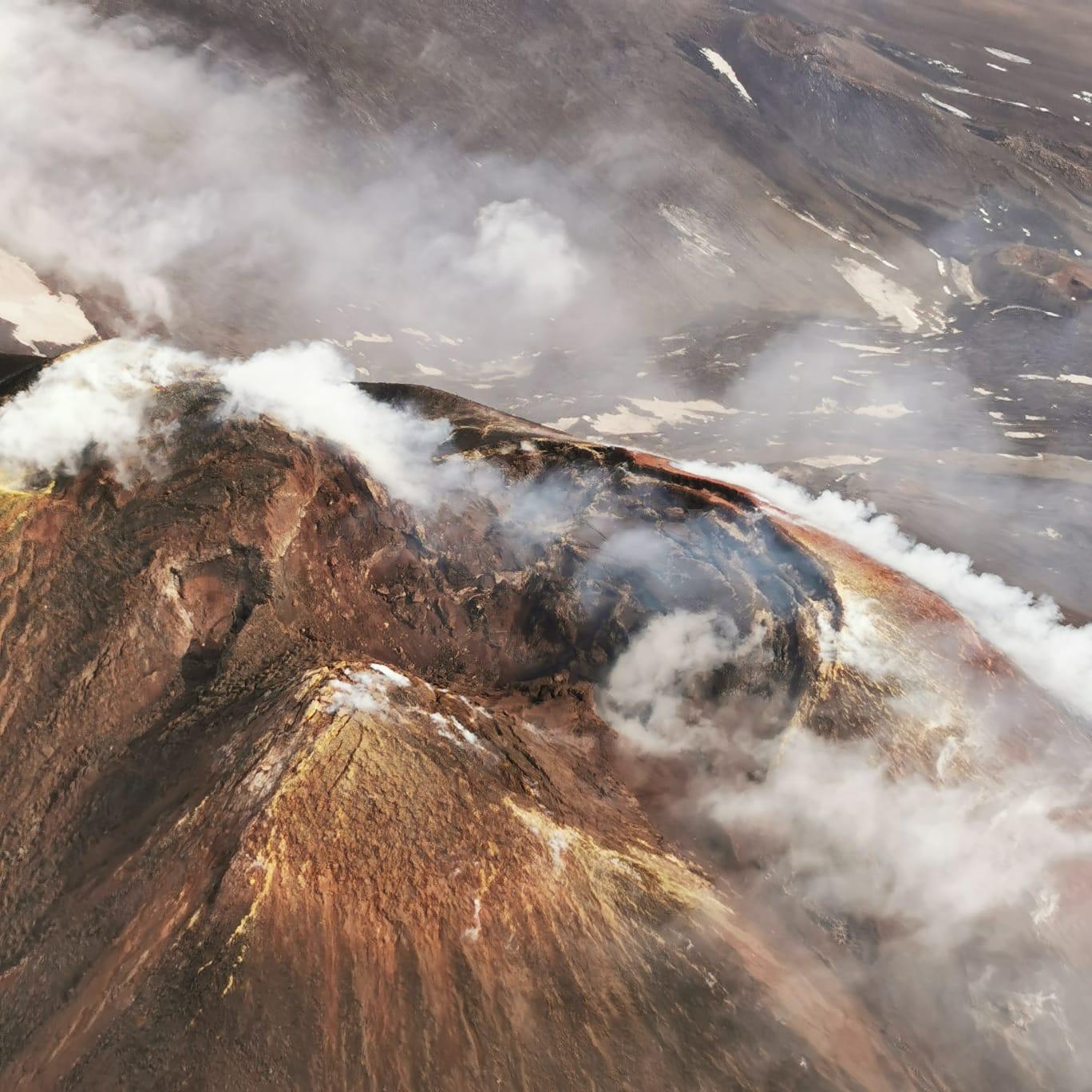 Aerial view of a steaming volcanic crater with rust-coloured slopes on Mount Etna