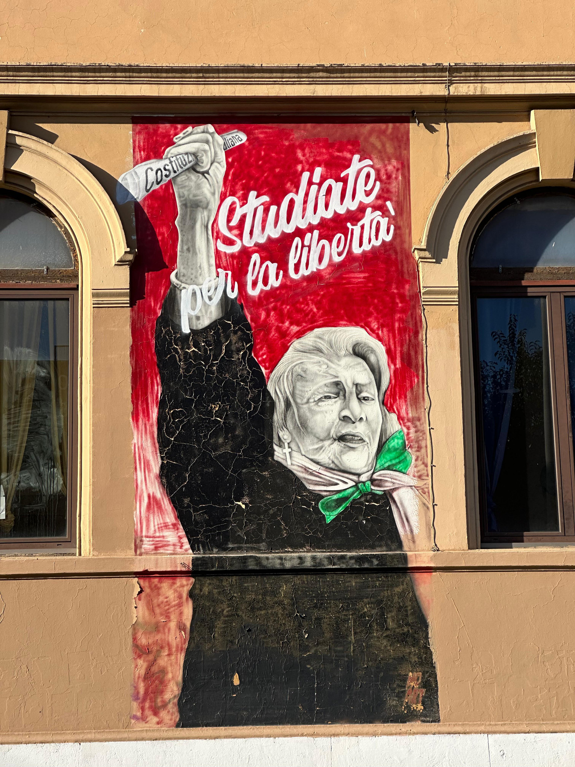 Red mural of an elderly woman with a green scarf holding a pencil, with the words 'Studiate per la libertà'