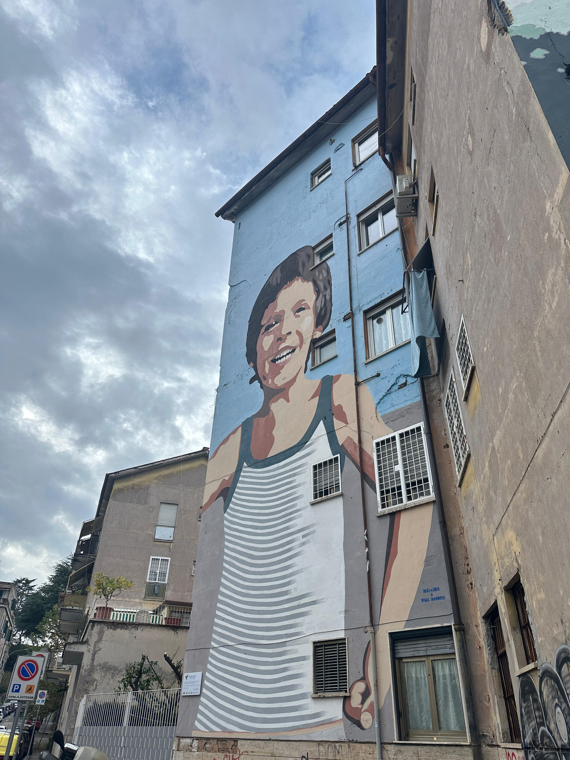 Tall mural of a smiling child in a striped top painted on the side of a blue apartment block