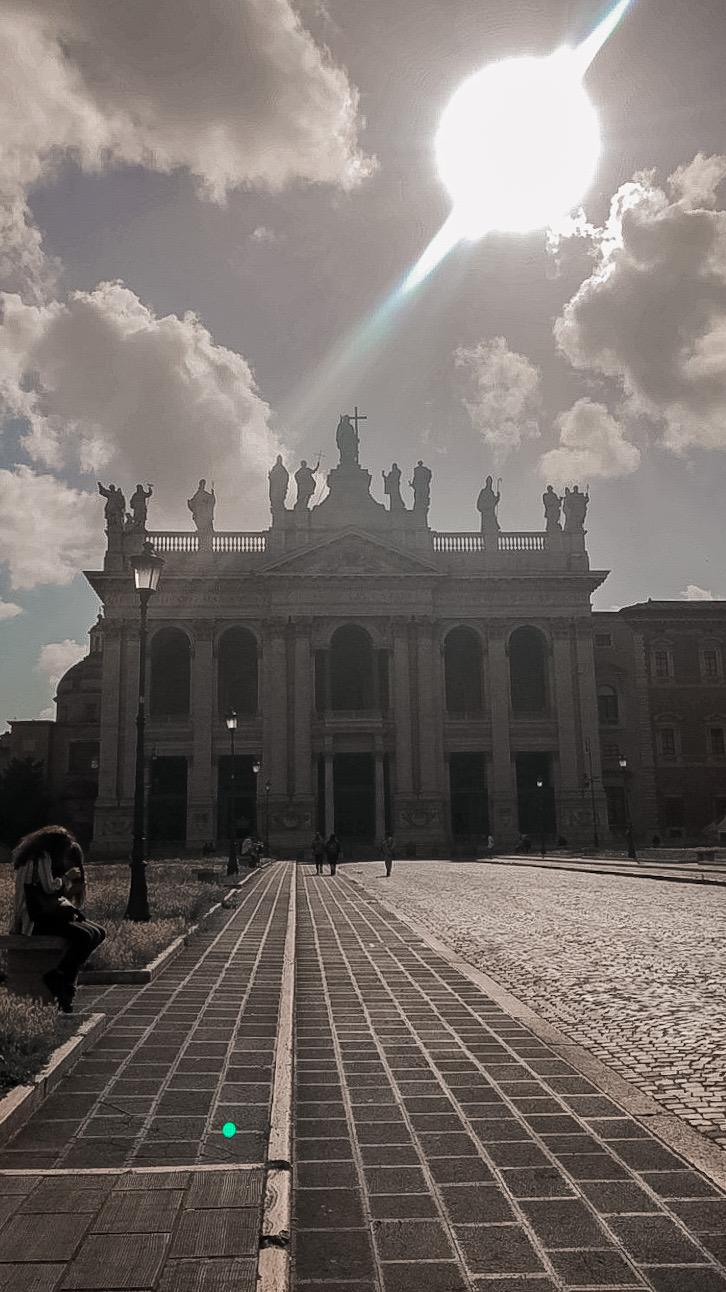 Backlit basilica facade crowned with statues, seen from a brick-paved square at sunset