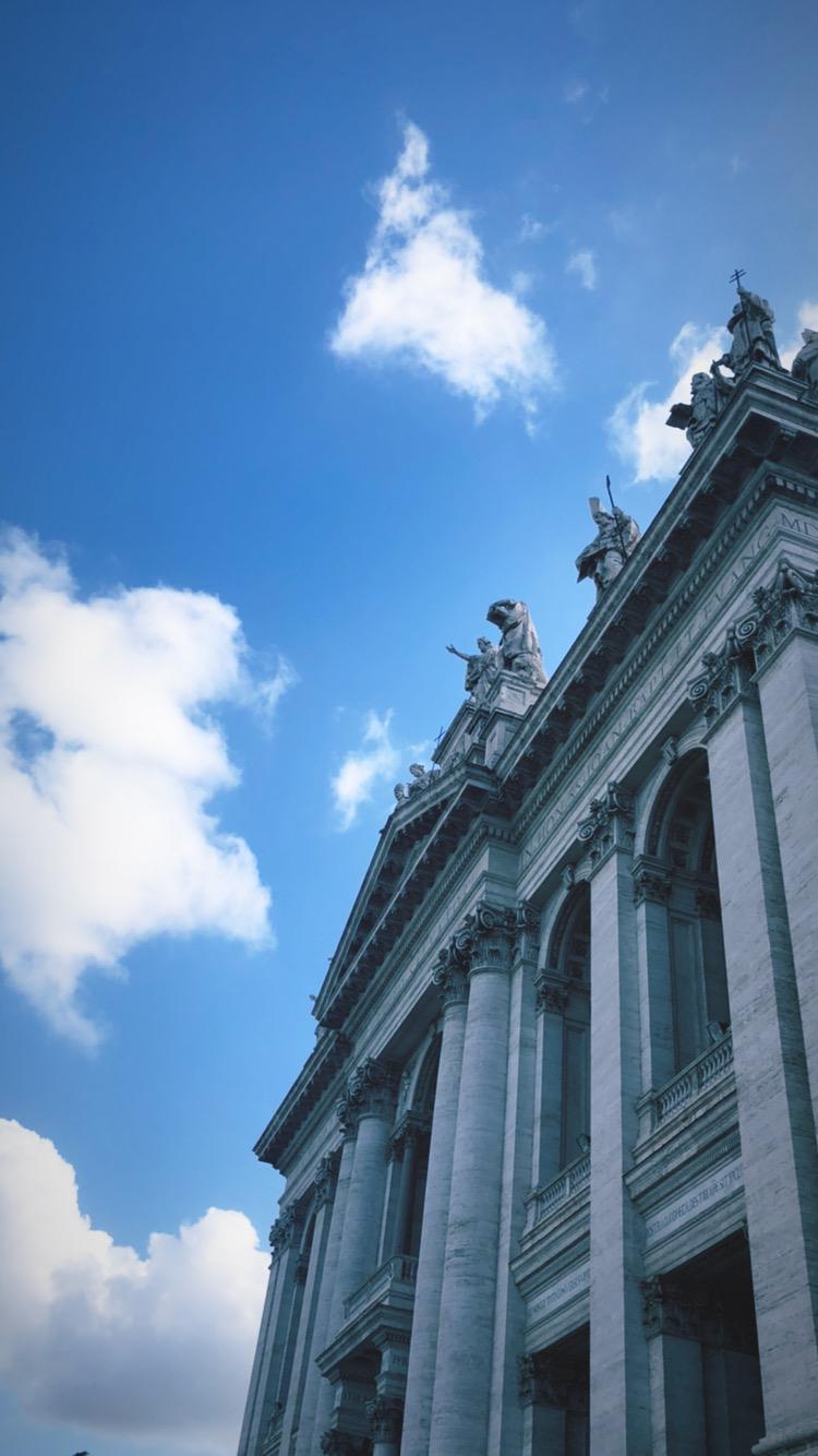 Statue-topped basilica facade and cornice rising against a blue sky with white clouds