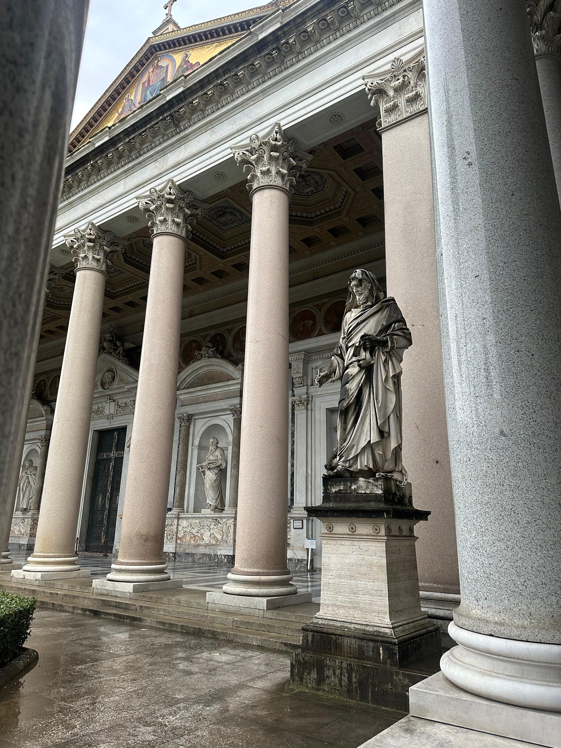 Rose granite columns and an apostle statue at the portico of a basilica on a rainy day