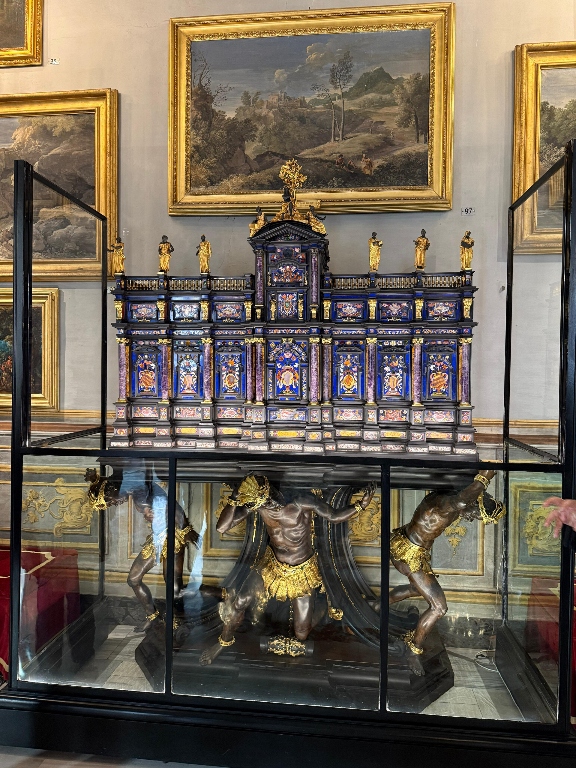 Ornate cabinet inlaid with blue lapis panels and gilded figures in a display case beneath landscape paintings