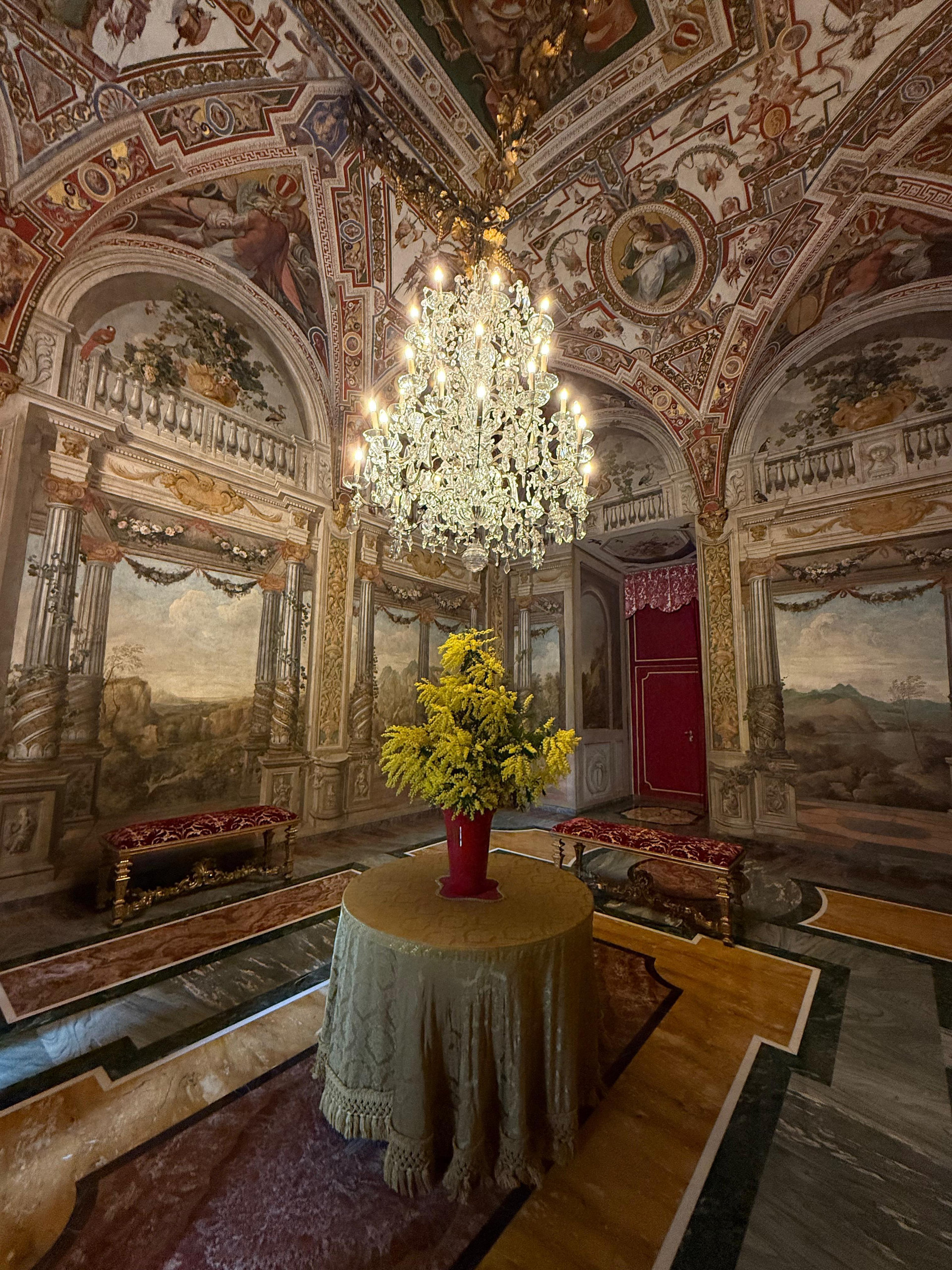 Frescoed salon with a chandelier and a round table holding yellow flowers in a red vase