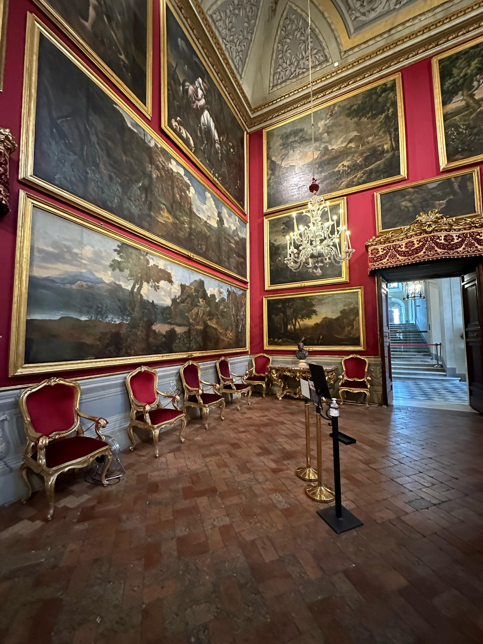 Red-walled gallery hung with large landscape paintings and gilded armchairs on a brick floor