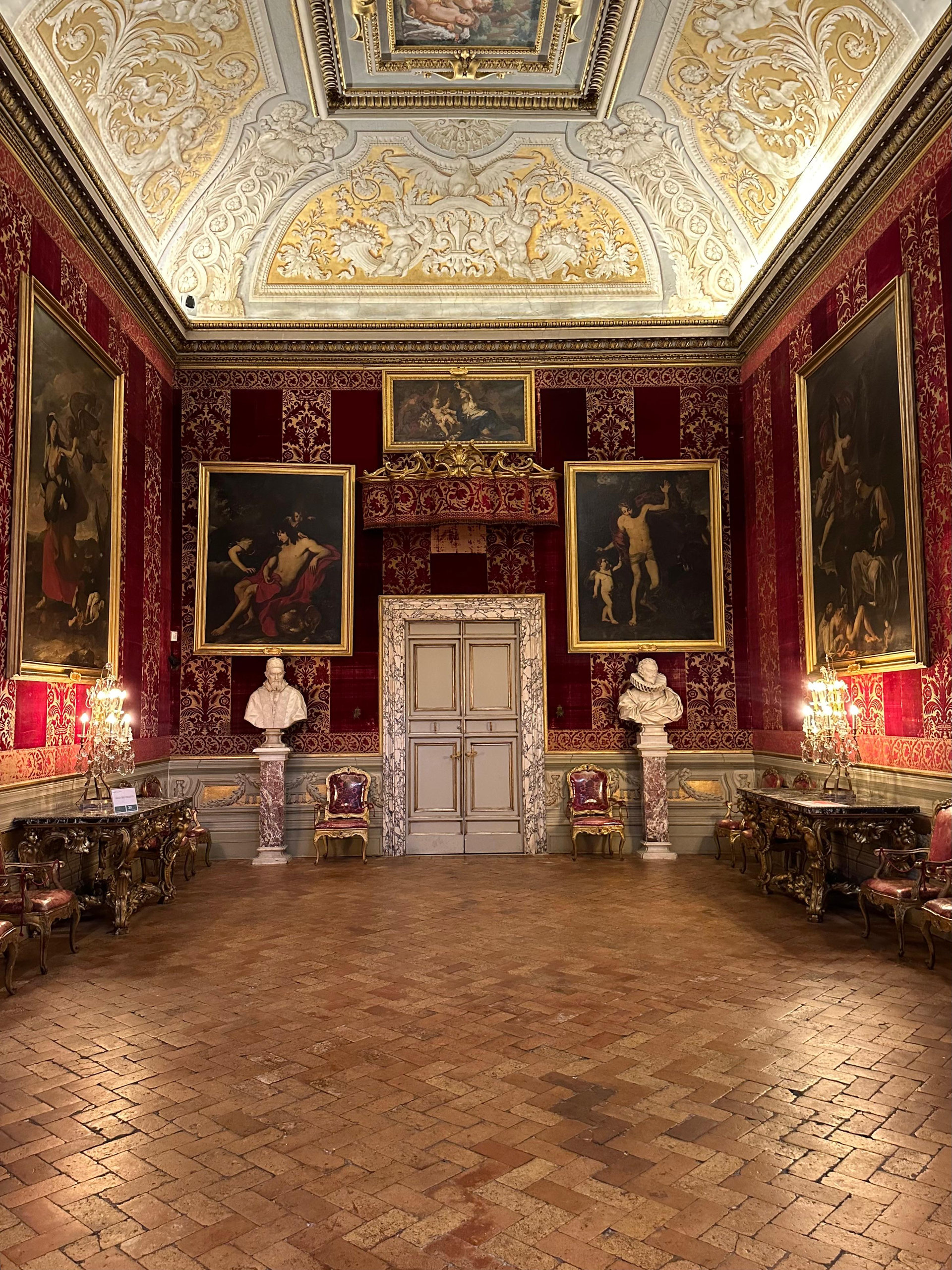 Red damask room with paintings, two marble busts and a stuccoed ceiling above a herringbone floor