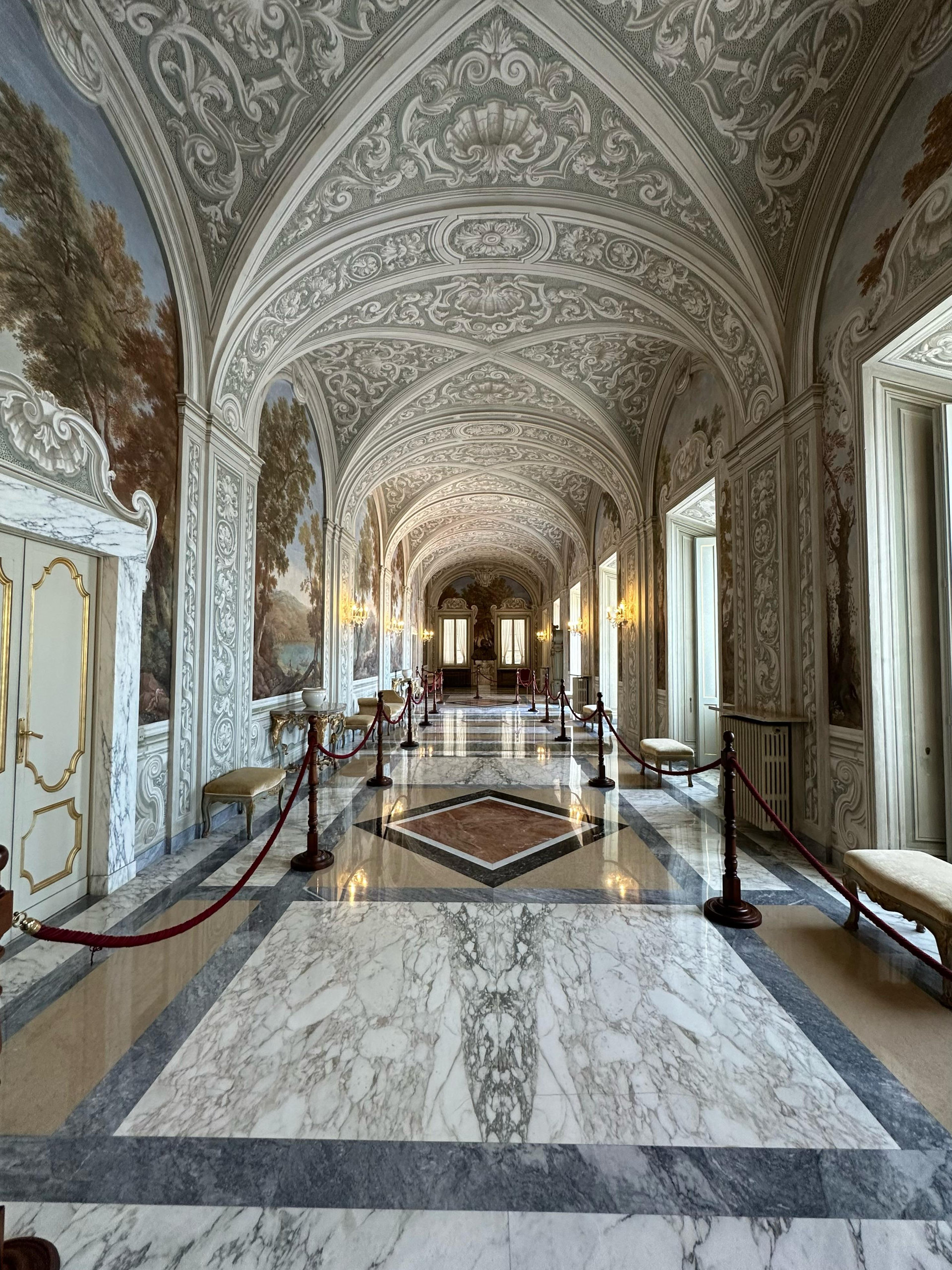 Vaulted stuccoed corridor with landscape frescoes and a marble floor between red ropes