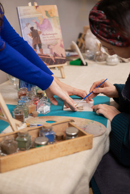 Participants painting at a table with small jars of pigments and an easel with an art book behind