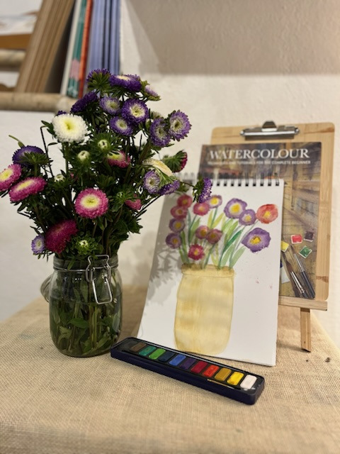 Watercolour sketch of a vase of flowers beside the real bouquet and a pan of paints