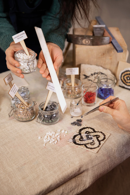 Hands arranging mosaic pieces with tweezers beside a finished knot-pattern mosaic and jars of coloured stones