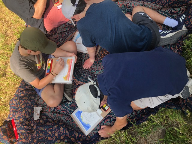 Young people painting watercolours while sitting on a patterned cloth on the grass