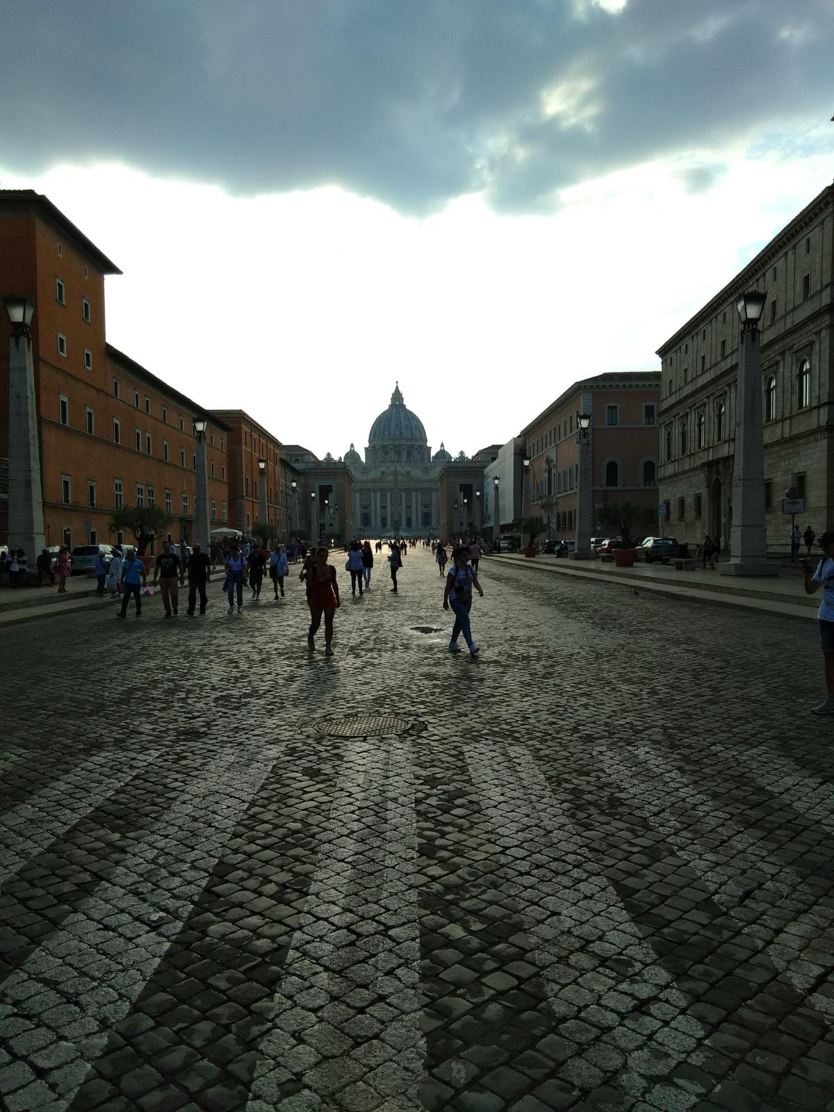 Walkers on a cobbled avenue leading towards the dome of Saint Peter's Basilica at dusk