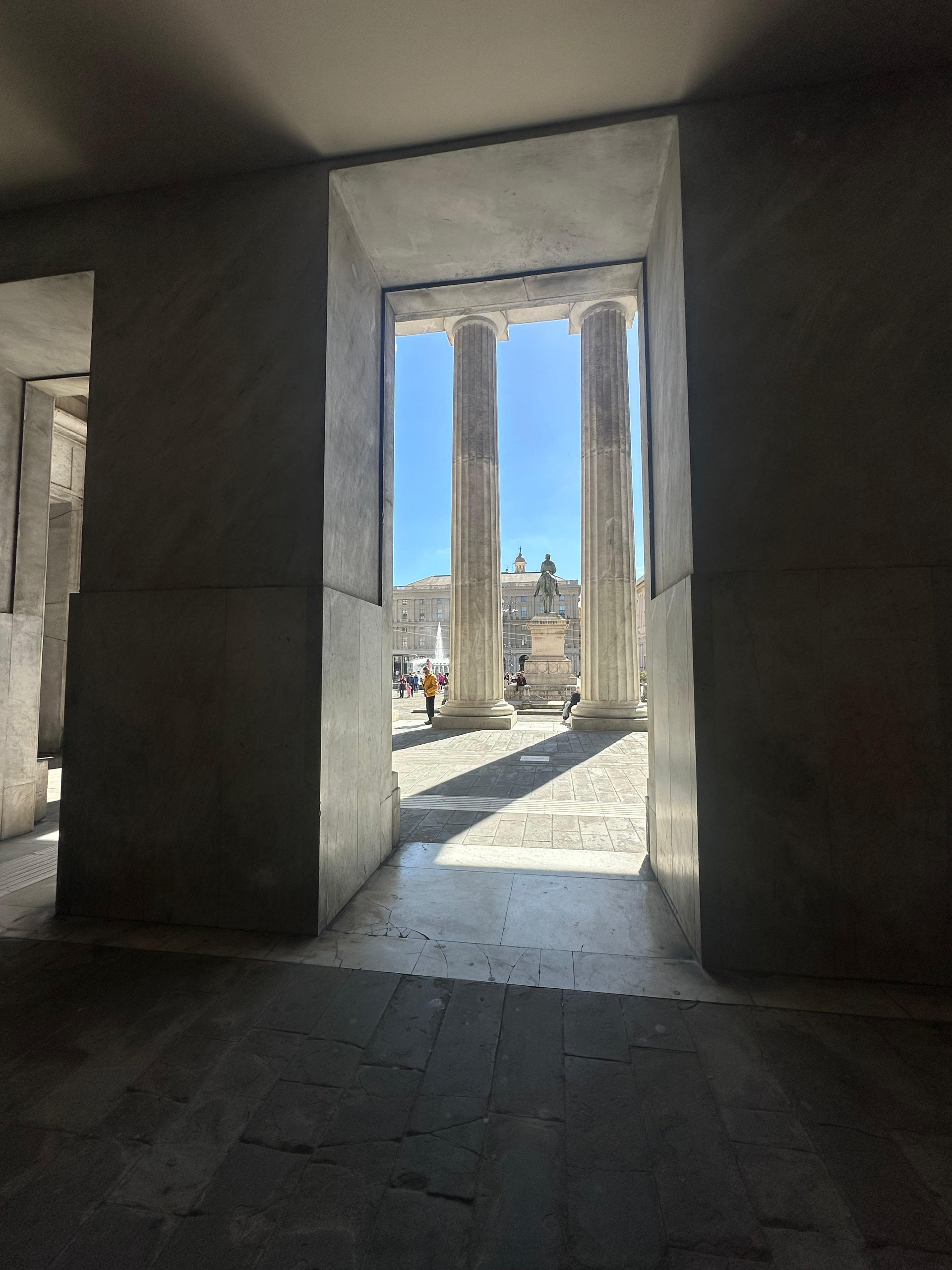 Marble doorway framing two stone columns and a statue in a sunlit square in Genoa
