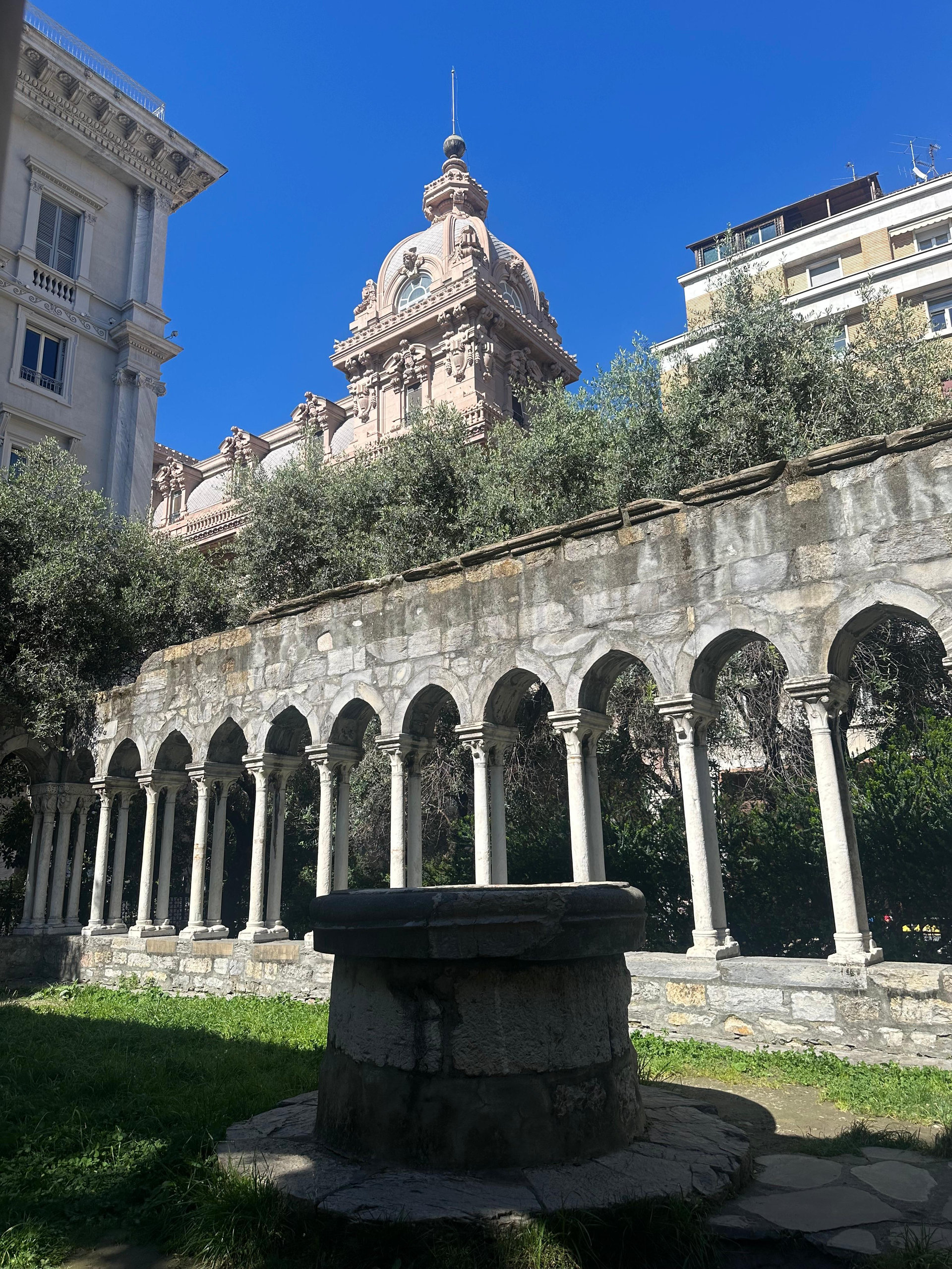 Medieval stone cloister with slender columns and a mossy well, a domed building rising behind