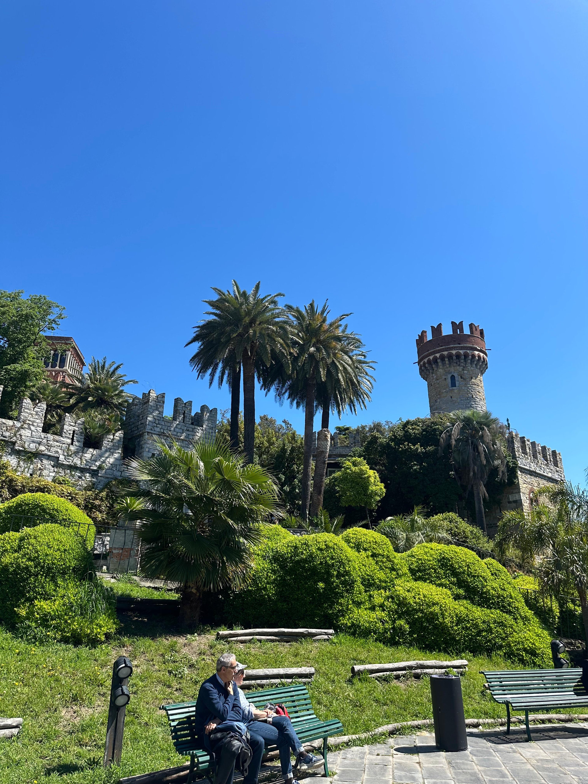 Palm trees and clipped hedges below a crenellated stone tower under a clear blue sky