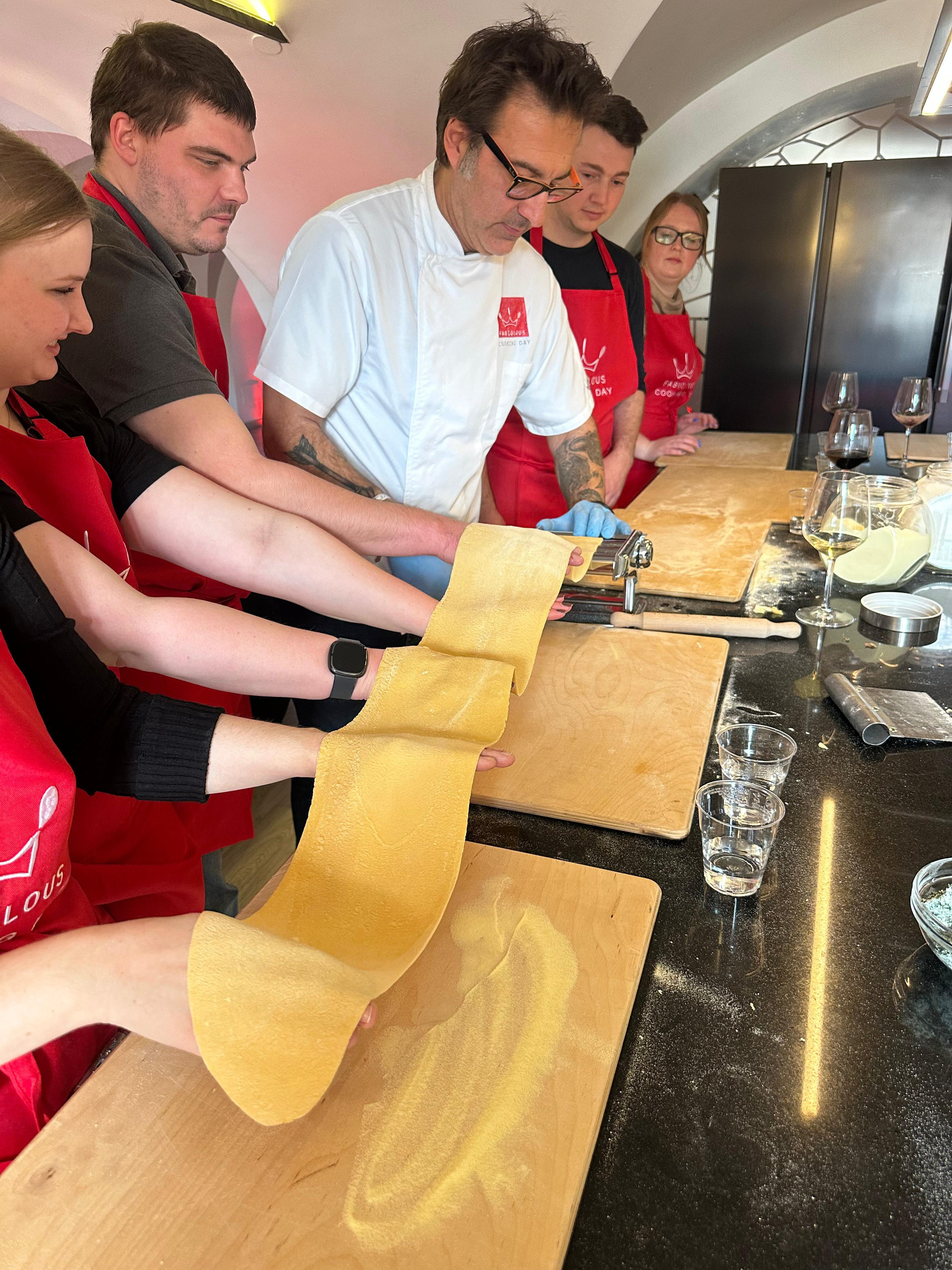 Group in red aprons stretching a sheet of fresh pasta dough with a chef during a cooking class