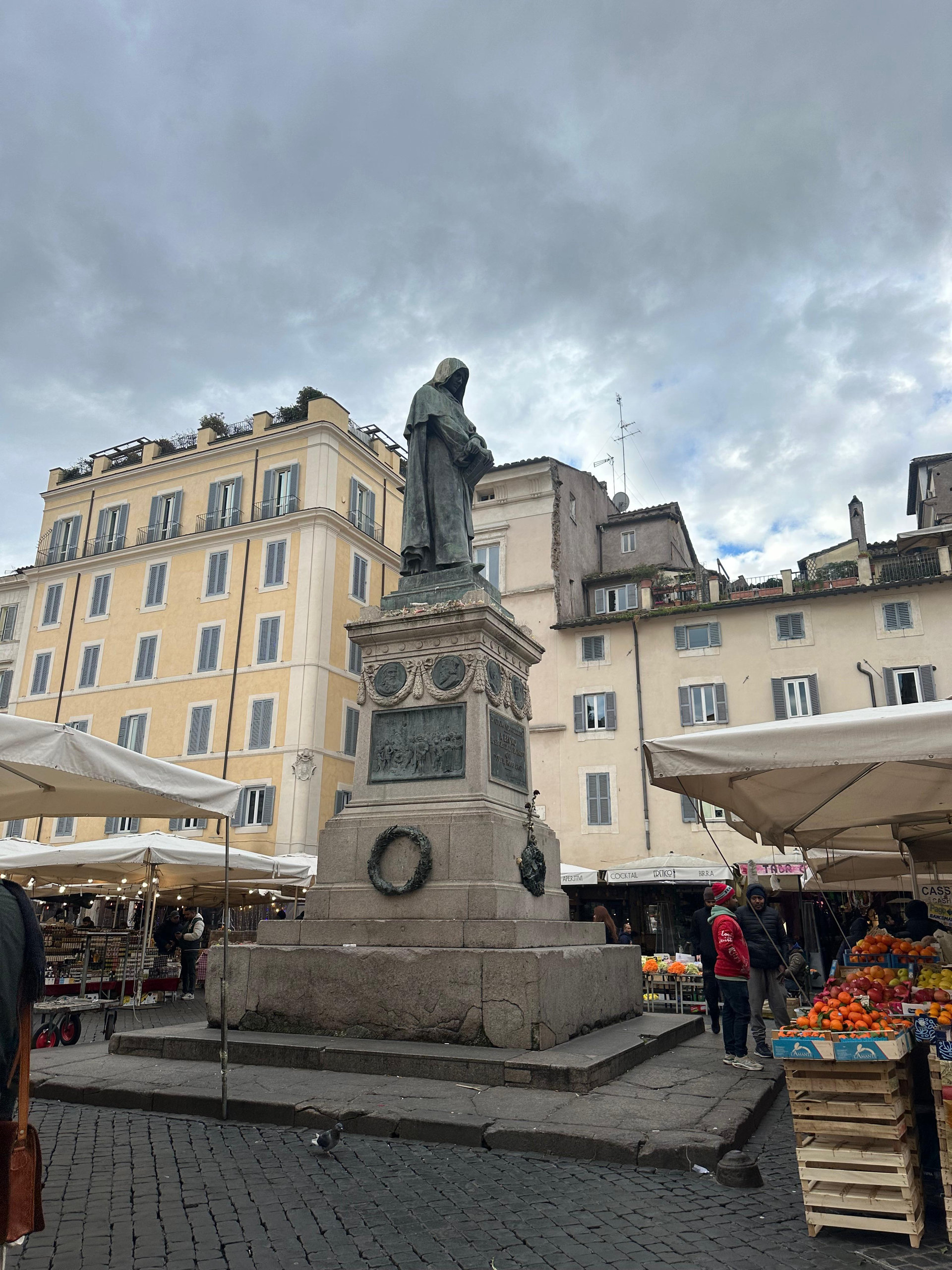 Hooded bronze statue on a stone pedestal in Campo de' Fiori, with market stalls and pastel buildings behind