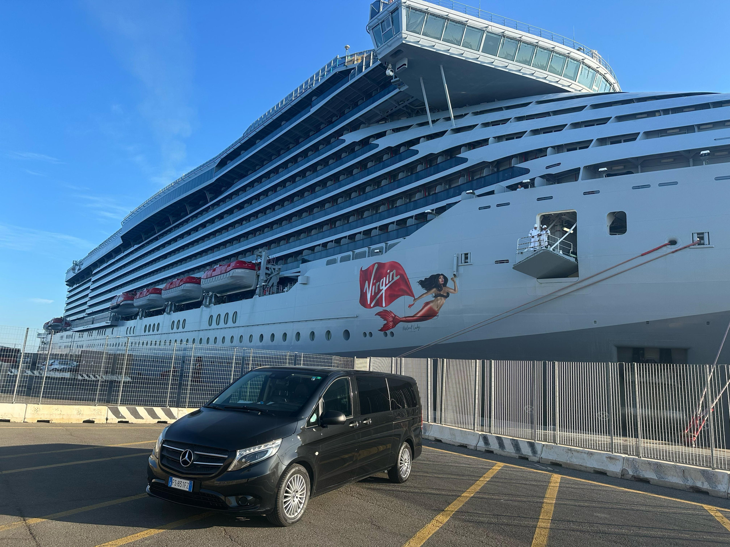 Black minivan waiting on the quay beside a large white cruise liner under a clear blue sky