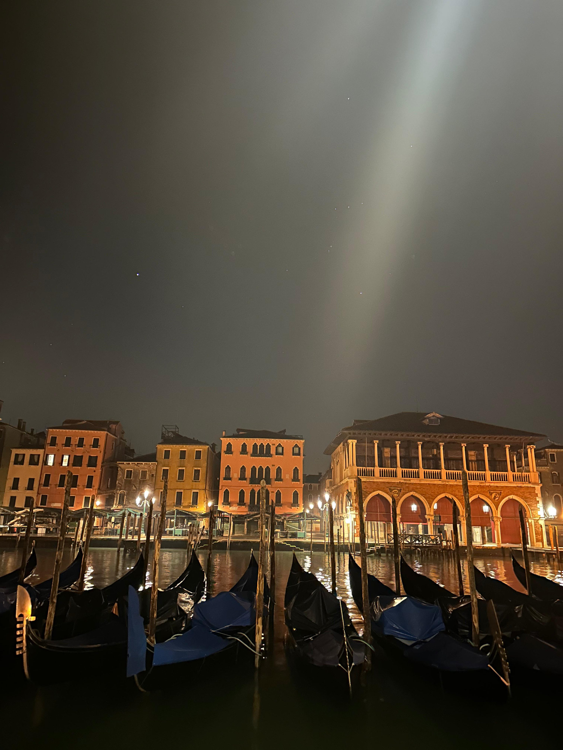 Moored gondolas by the Grand Canal at night, with a beam of light in the dark sky and lit palaces behind