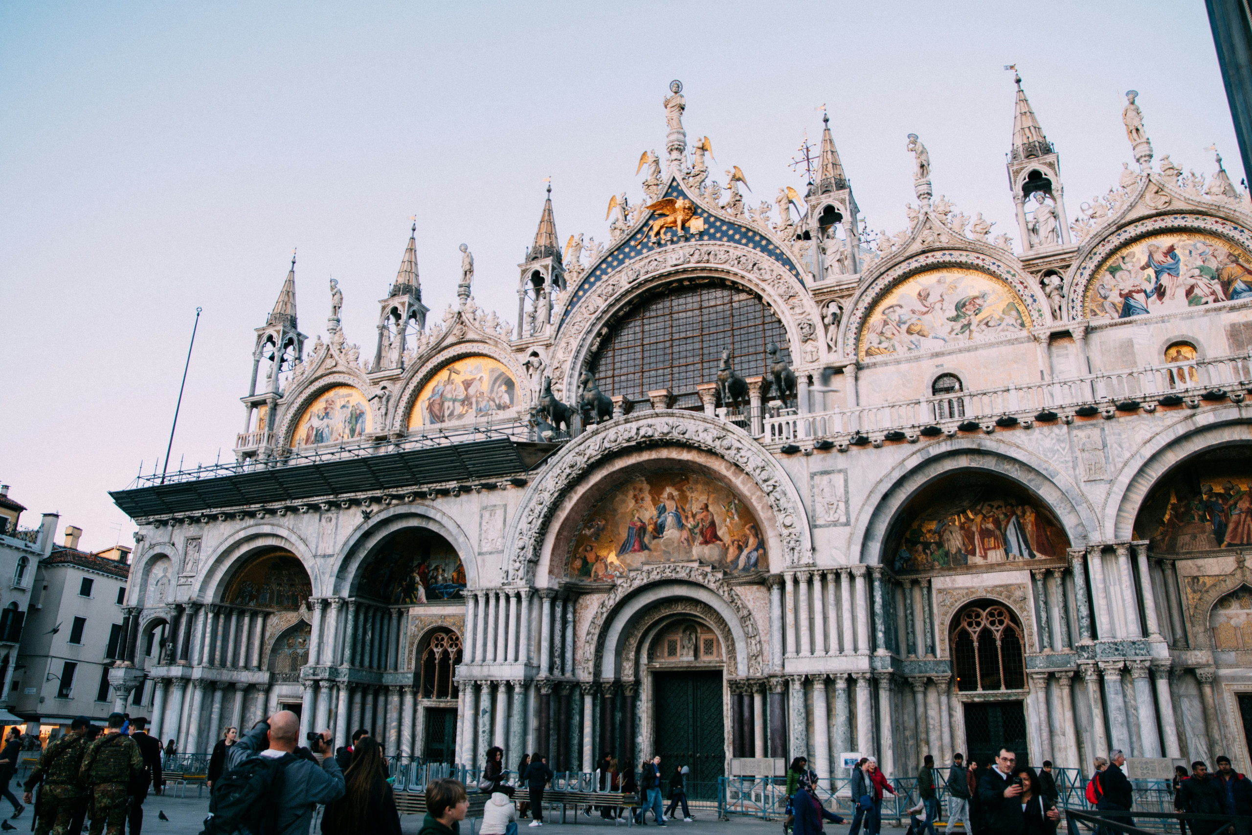 Facade of St Mark's Basilica with its golden mosaics, arches and pinnacles, with visitors in the square below