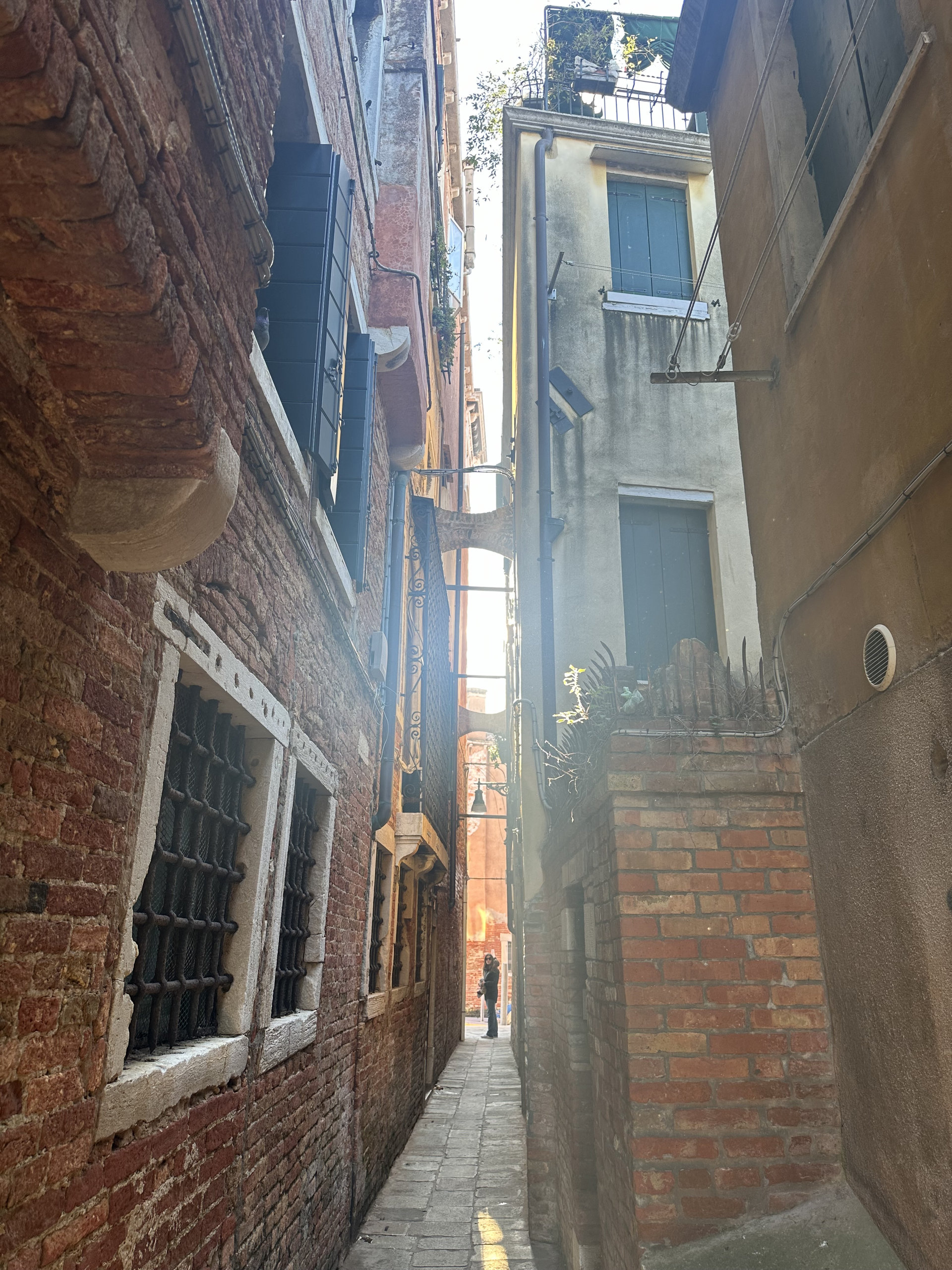 A narrow Venetian alley between red brick walls with barred windows and a walker in the distance
