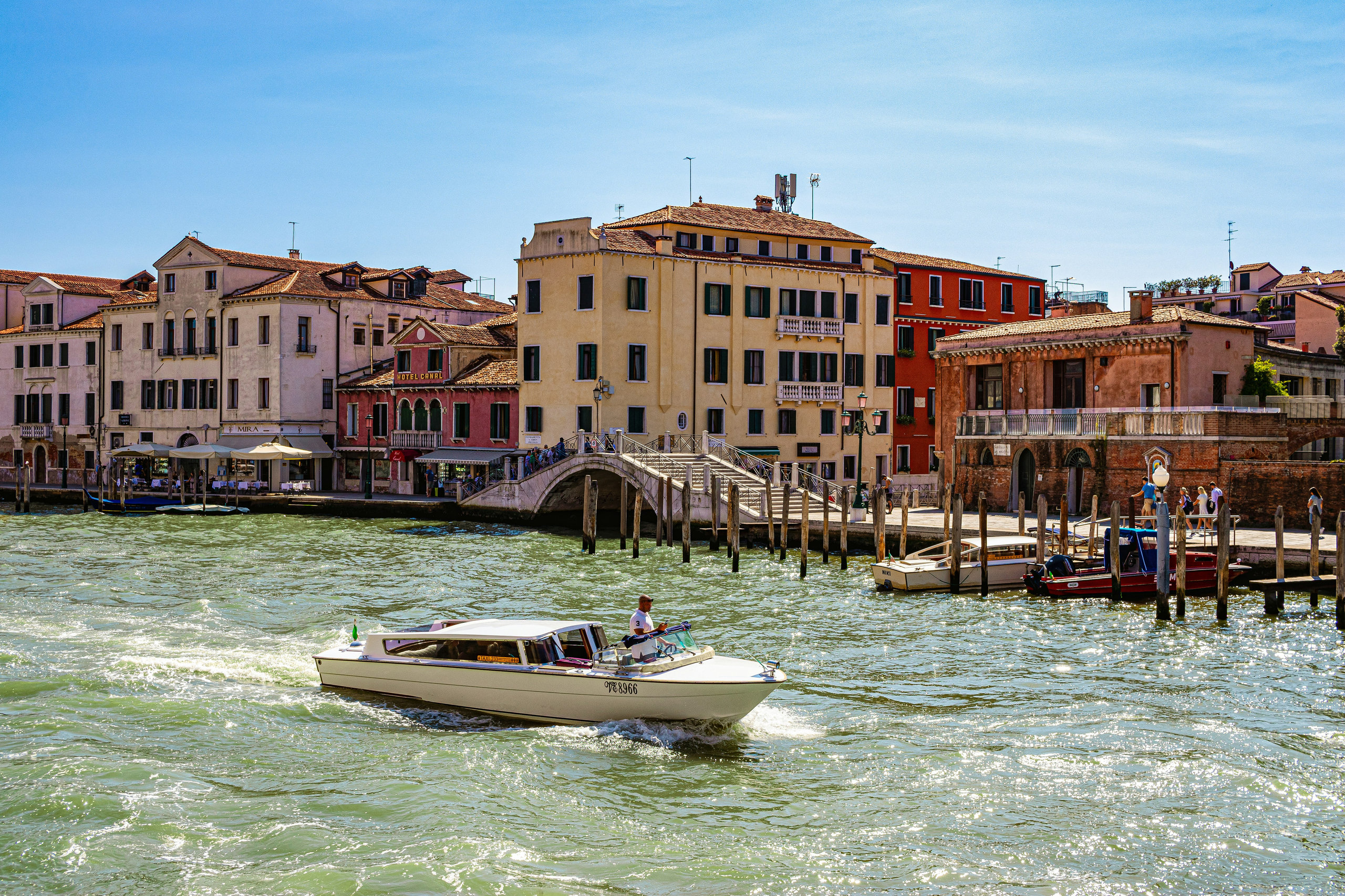 Motorboat crossing a sparkling Venetian canal in front of an arched footbridge and ochre buildings