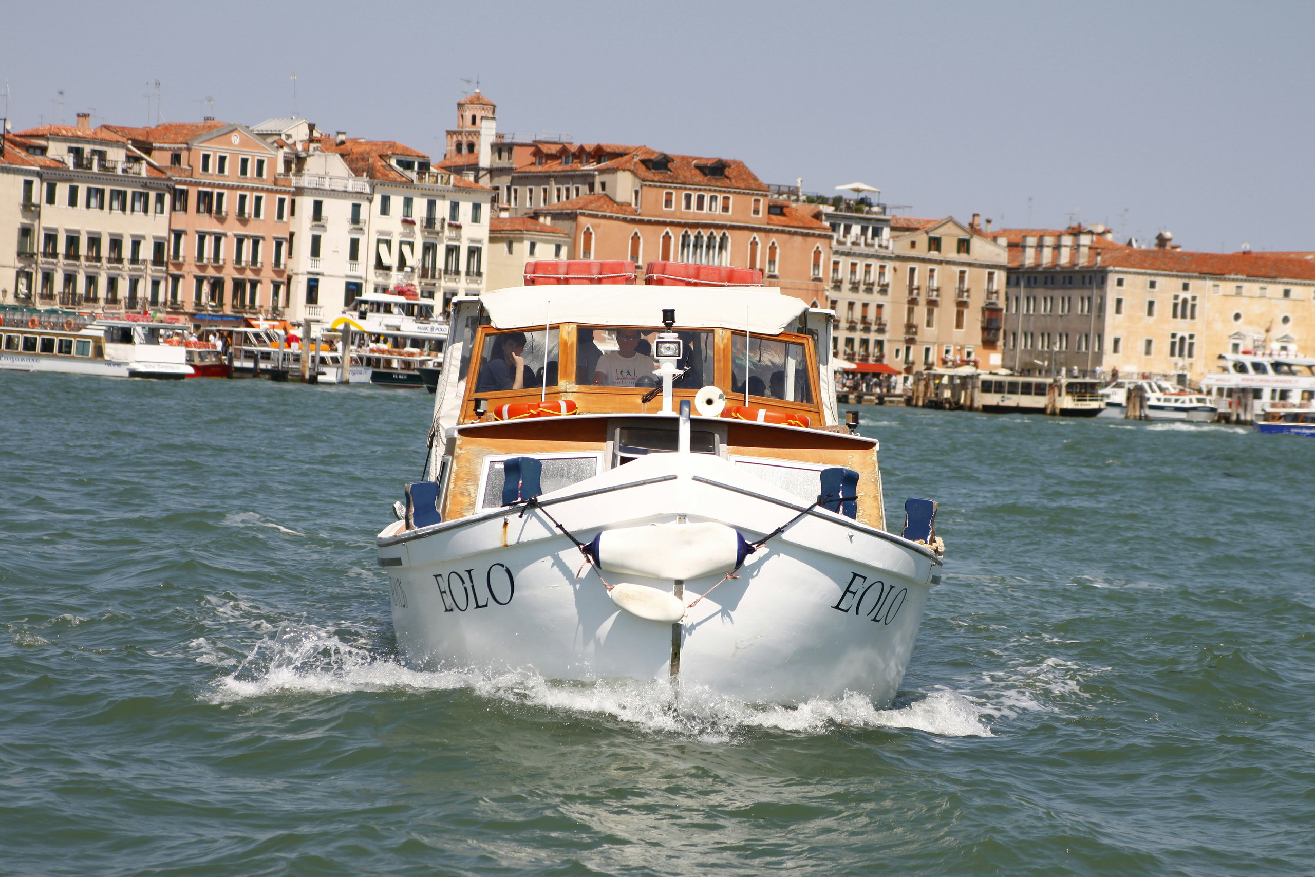 White wooden water taxi heading towards the camera on the Venetian lagoon, with waterfront buildings behind