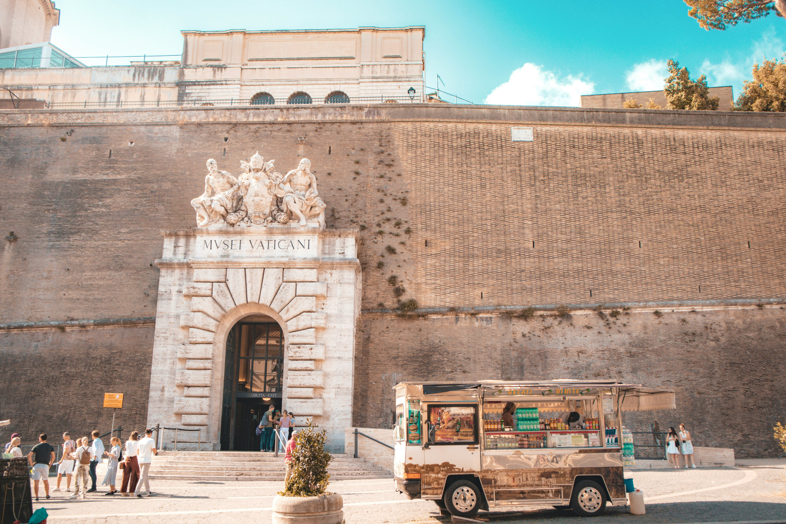 Vatican Museums entrance in a high brick wall, with a sculpted pediment, visitors queuing and a food truck outside