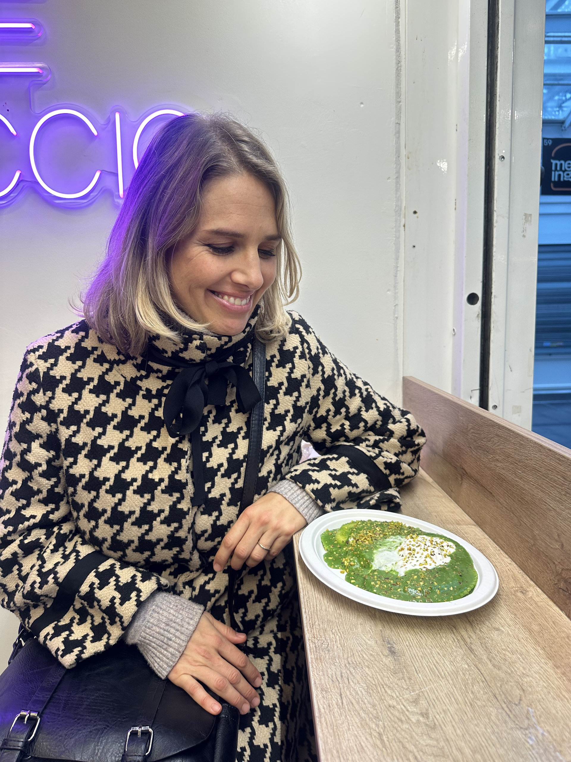 Smiling traveller looking at a plate of green sauce topped with cream, seated beside a neon sign