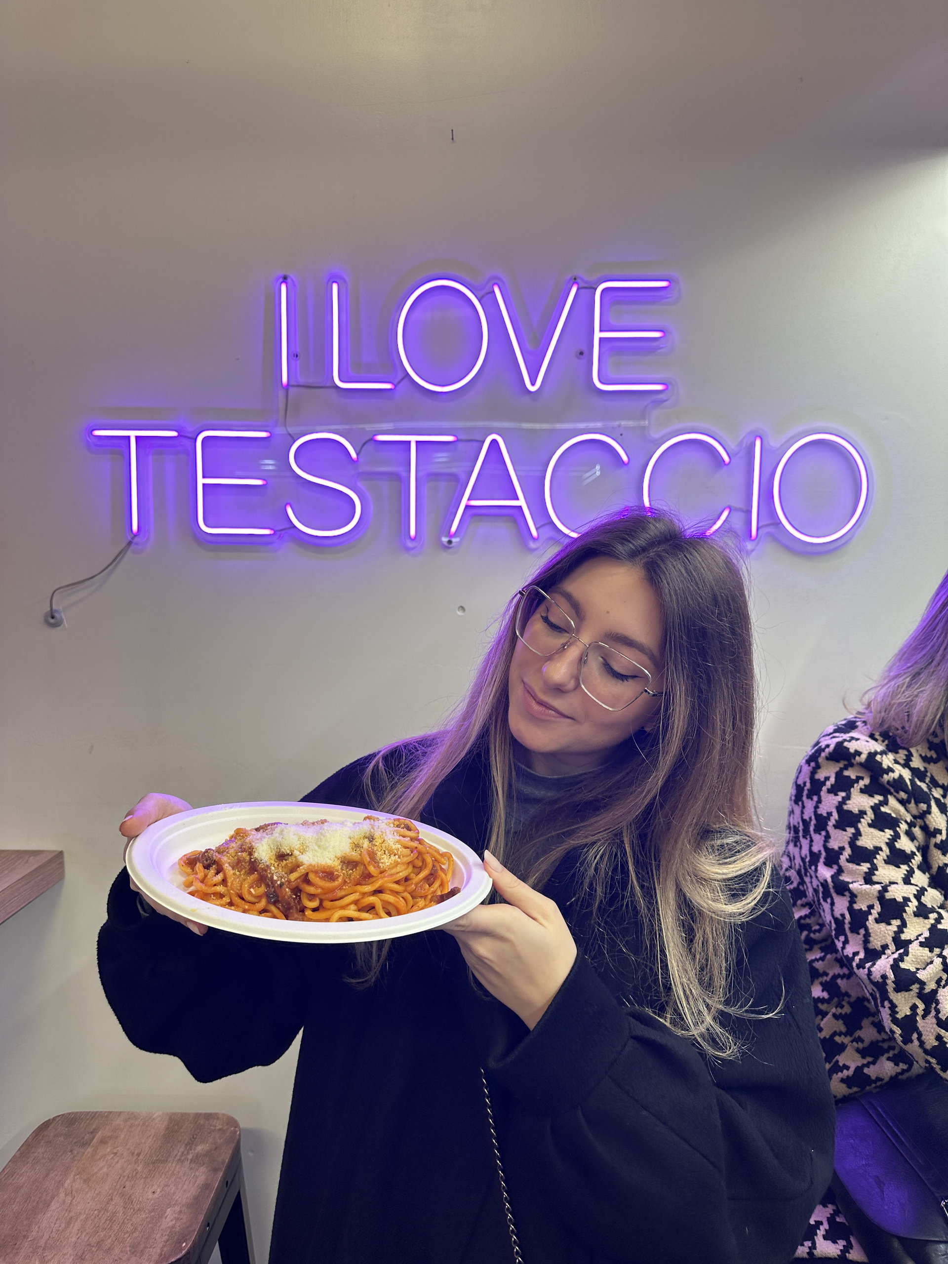 Traveller holding a plate of pasta in tomato sauce under a purple neon sign reading I love Testaccio