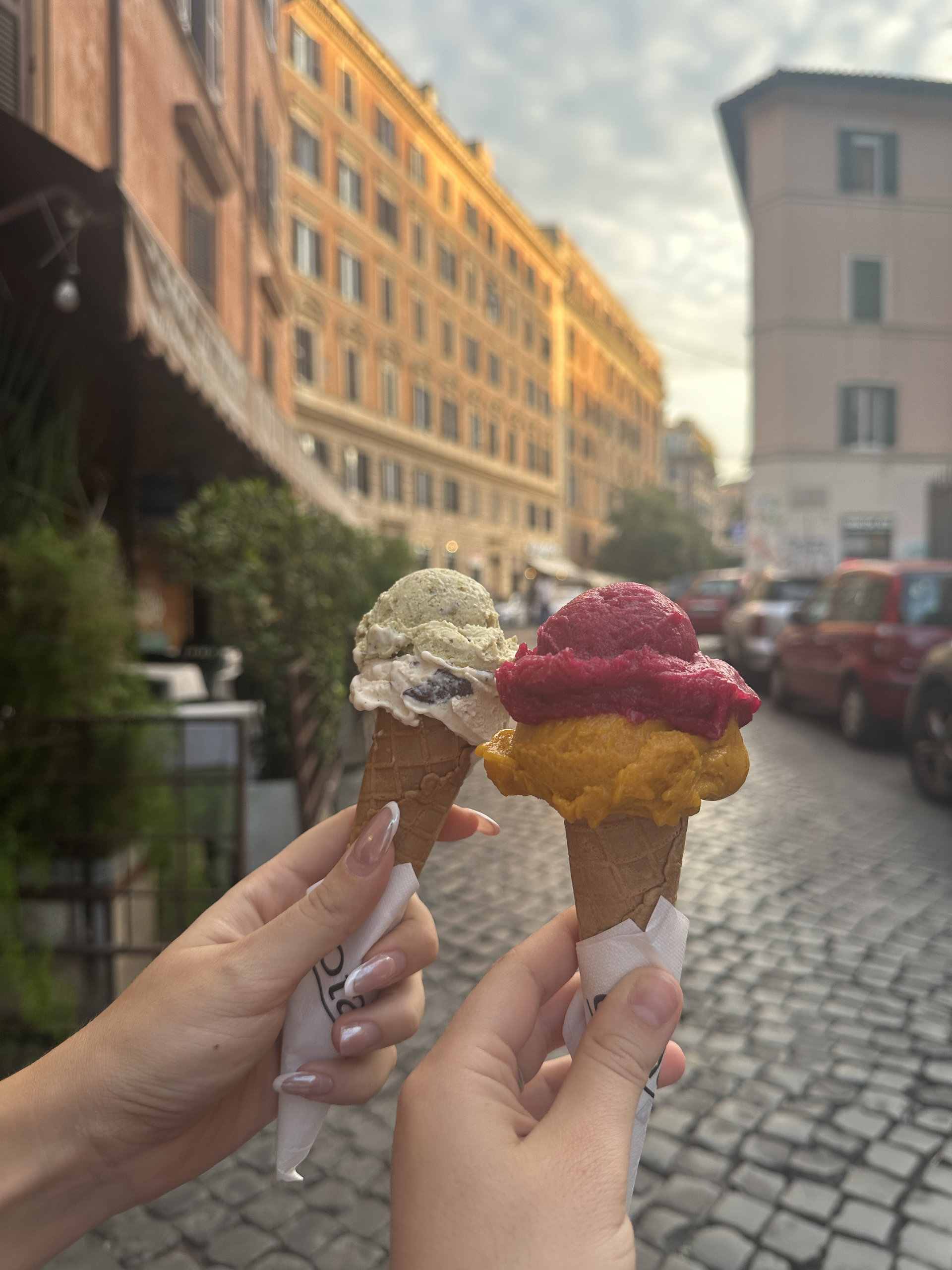Two hands holding gelato cones in different flavours on a cobbled Roman street