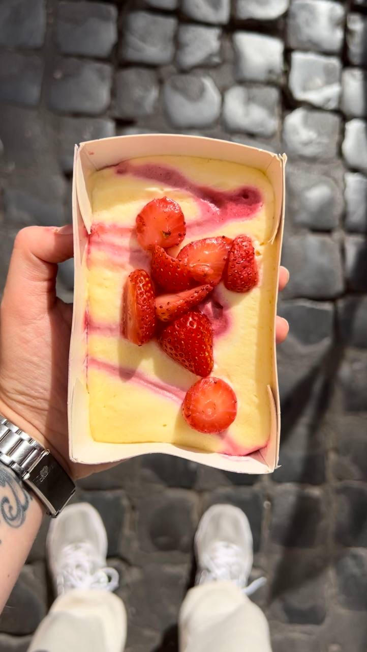 Cardboard tray of creamy dessert topped with fresh strawberries, held above cobblestones