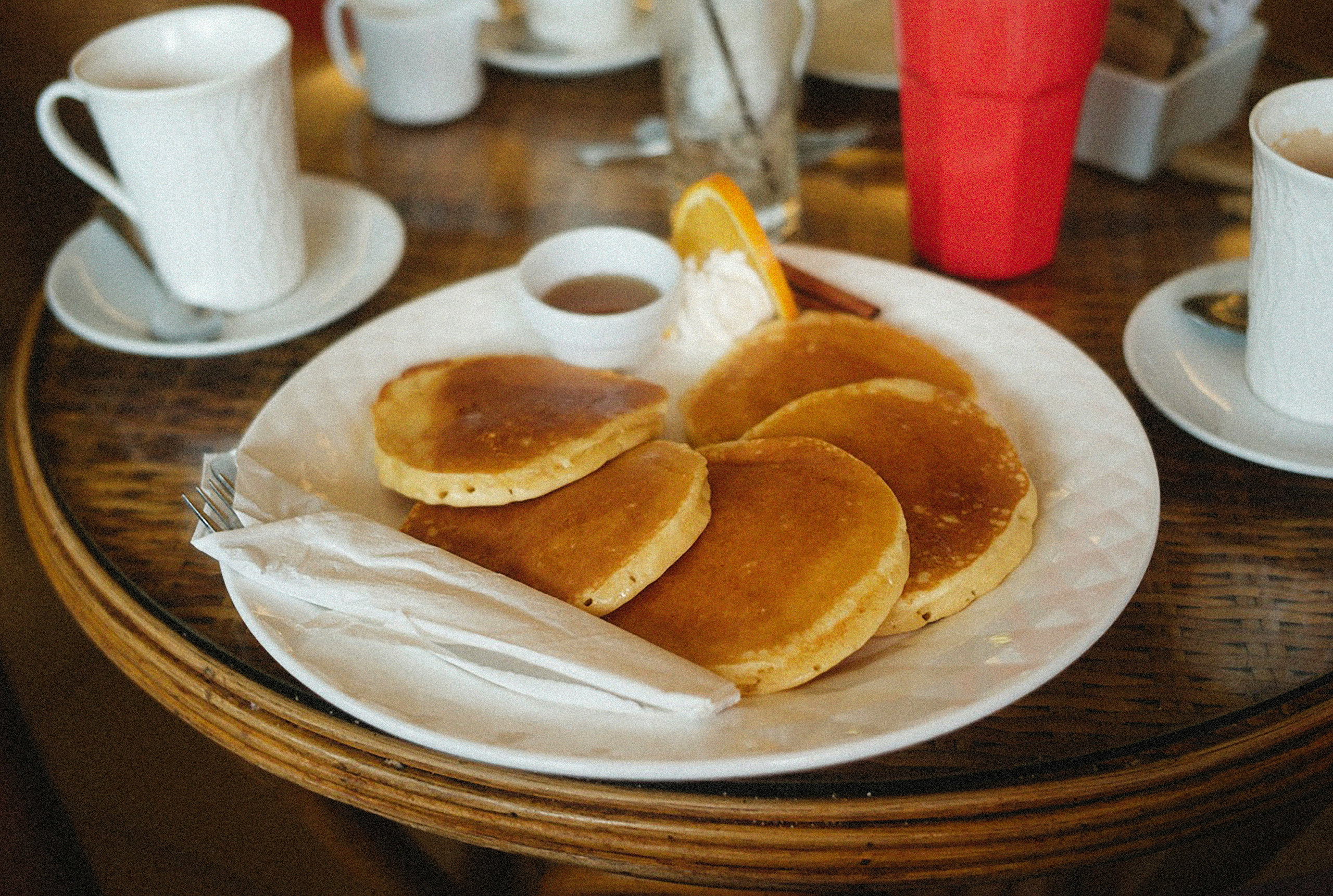 Plate of golden pancakes with syrup and an orange slice, served with cups of coffee