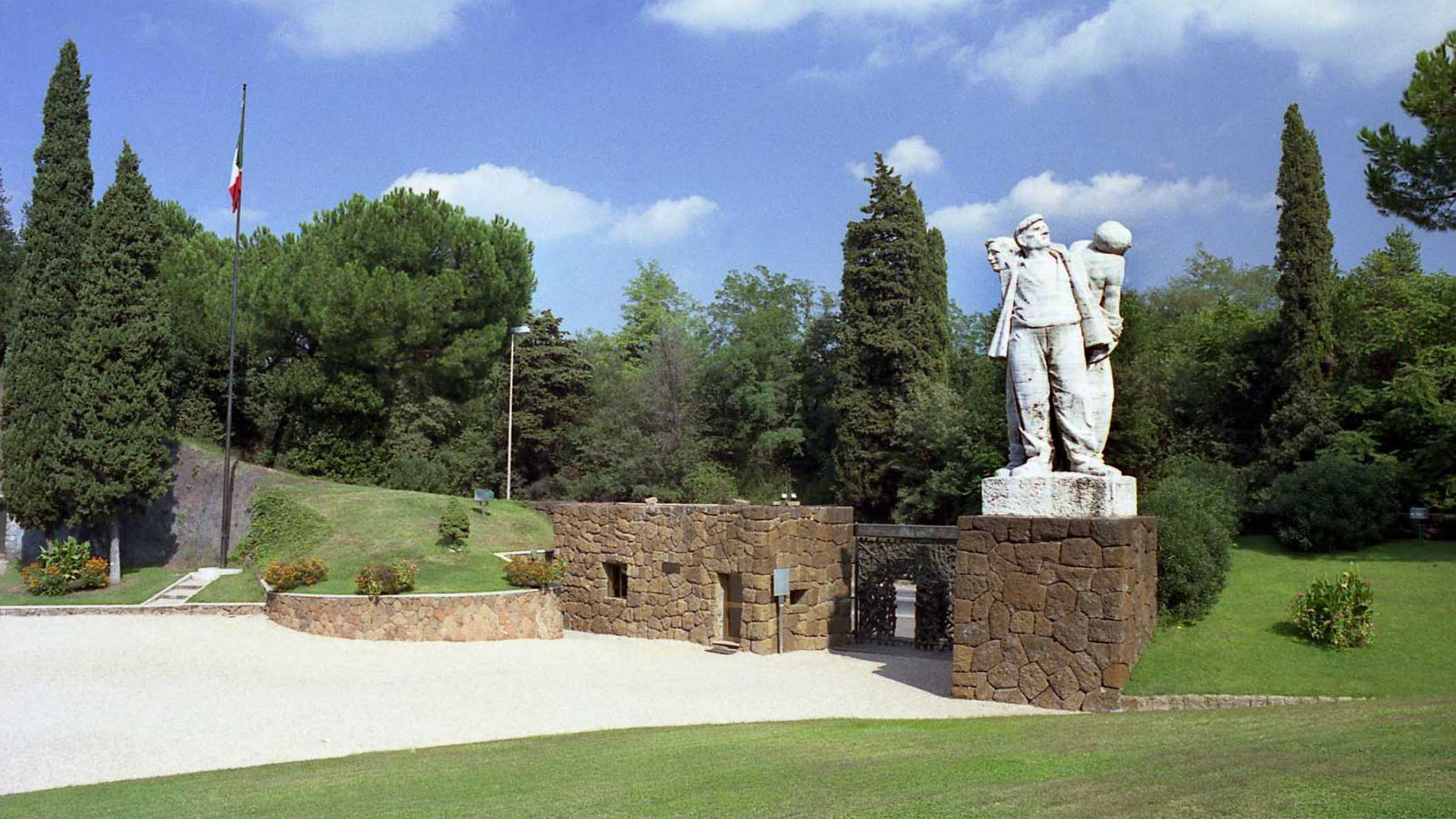 Memorial sculpture above a stone gateway, with a flag and cypress trees on a grassy slope