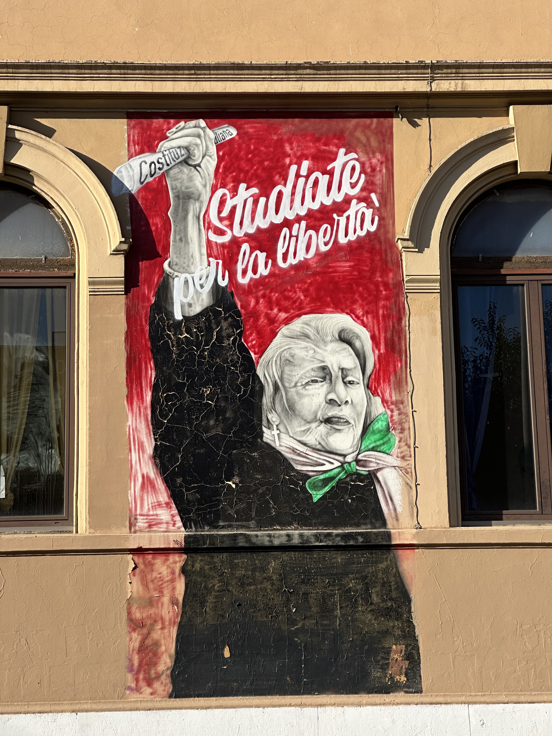 Red mural of an elderly woman with a green scarf raising a pencil, with the words 'Studiate per la libertà'