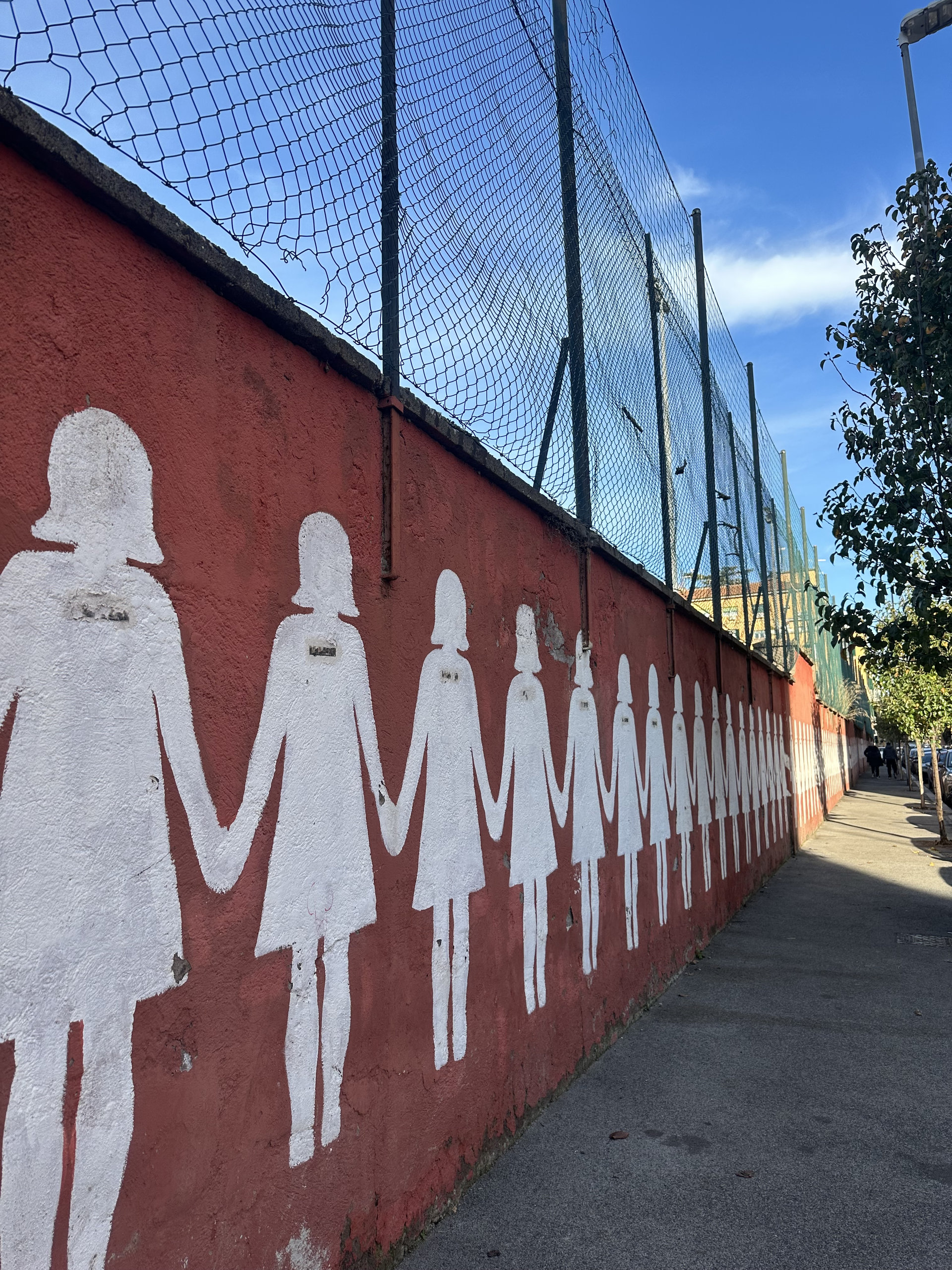 Row of white painted figures holding hands along a red wall beside a pavement