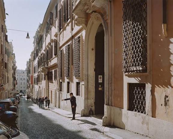 A cobbled Roman street lined with historic palaces and a man in a dark suit gesturing
