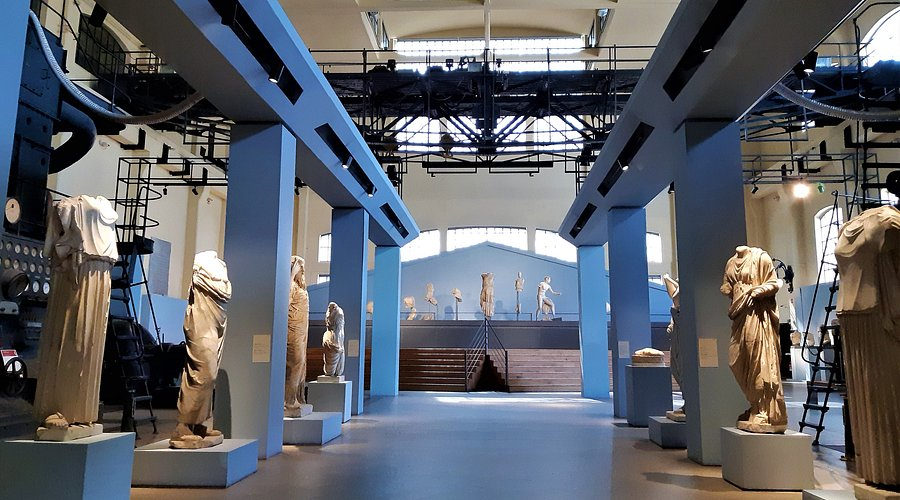 Draped Roman statues lined up along the industrial hall between blue steel columns