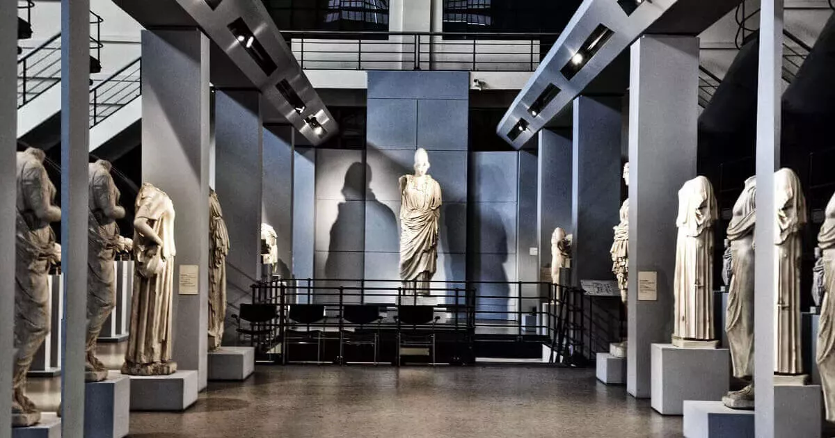 Marble statue lit against a pale wall, with rows of headless statues on plinths under steel girders