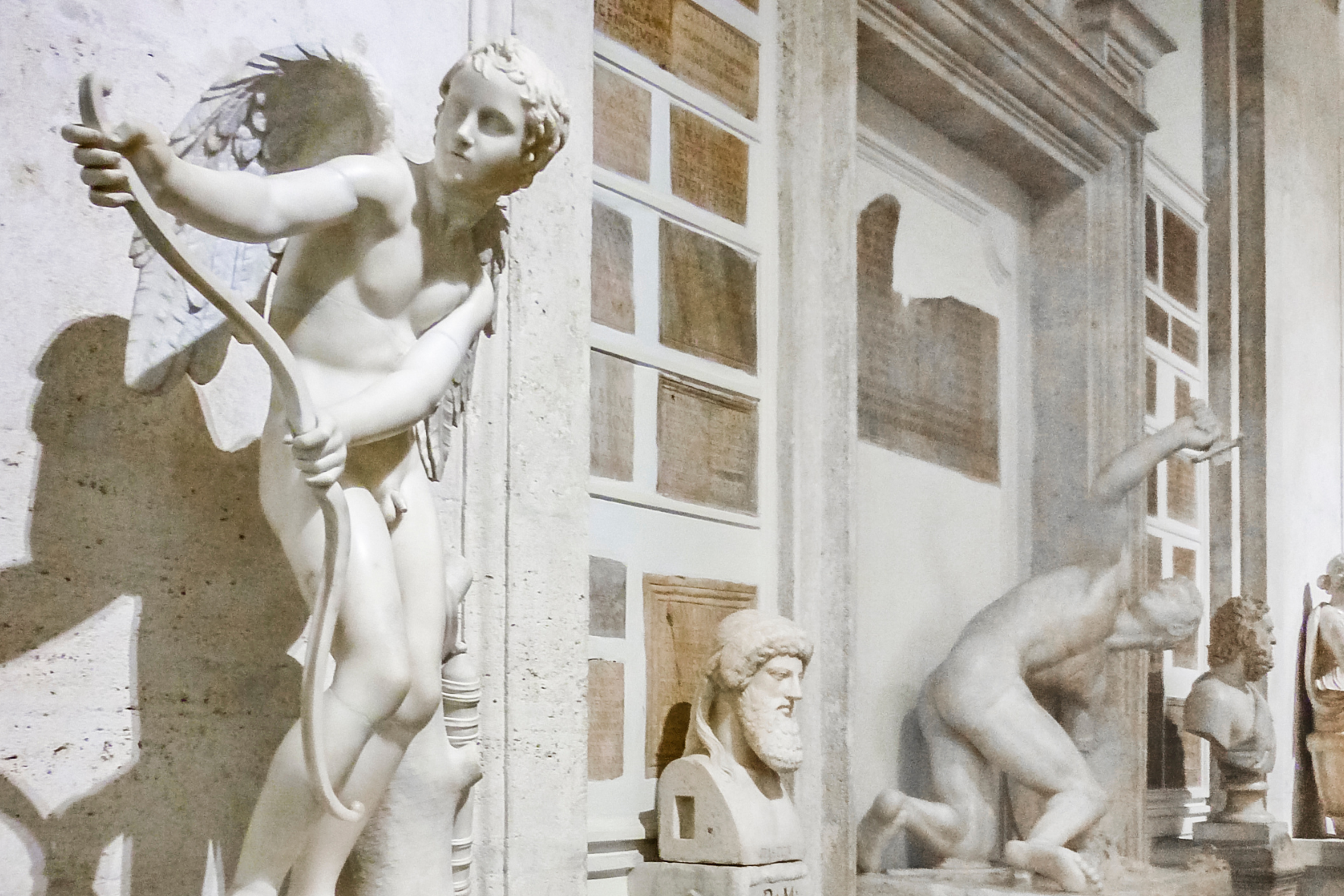 Marble statue of Cupid drawing a bow, beside ancient busts and statues in a museum hall