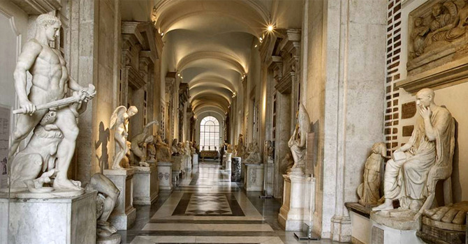Long vaulted gallery of ancient statues, including a muscular figure with a club, leading to an arched window