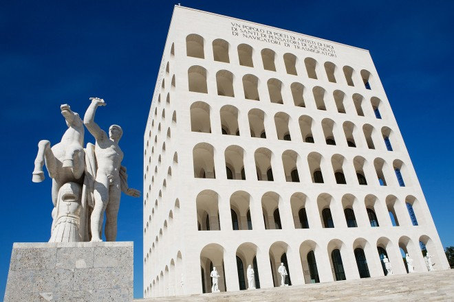 Square white building of stacked arches and a statue group, seen from below against a deep blue sky