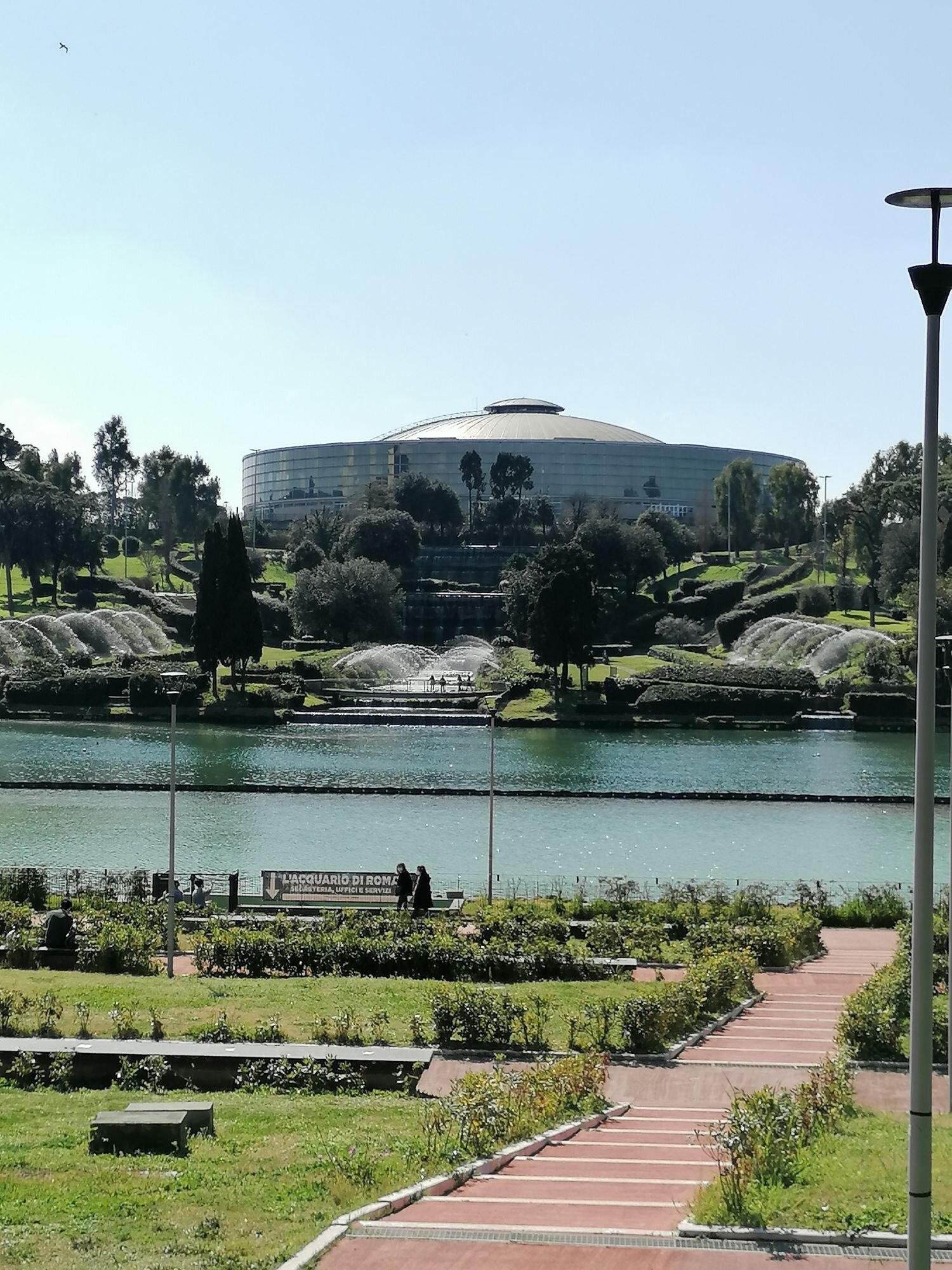 Park lake with fountains and a large domed arena behind the trees, with a couple strolling the shore