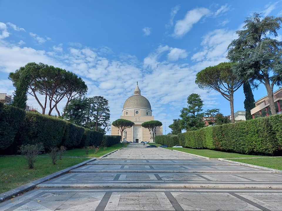 Domed basilica at the end of a paved esplanade framed by clipped hedges and umbrella pines