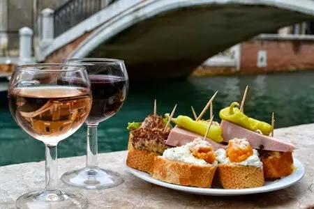 Glasses of wine and a plate of cicchetti on a canal-side ledge with a stone bridge behind