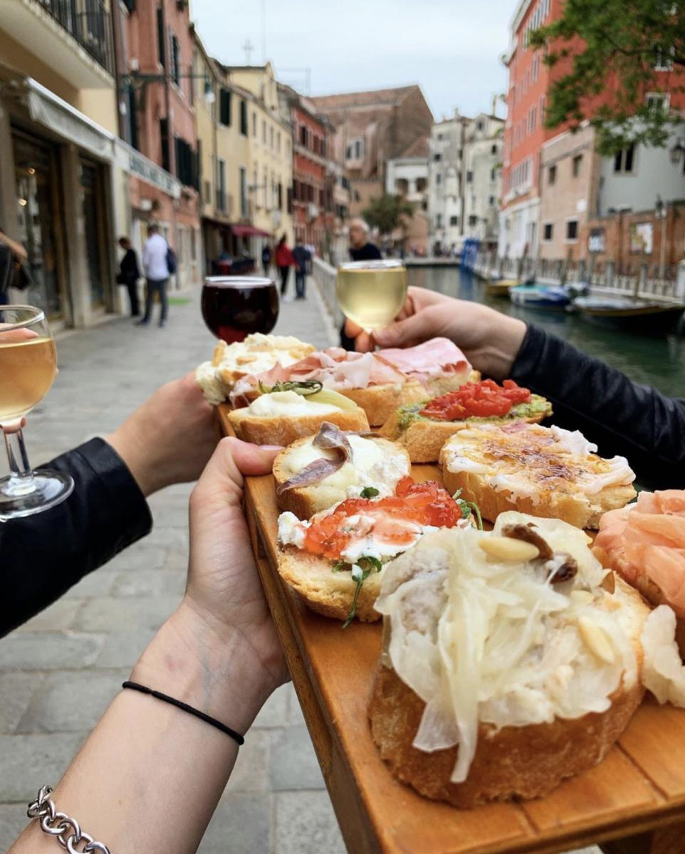 Hands sharing a wooden board of cicchetti and glasses of wine along a Venetian canal