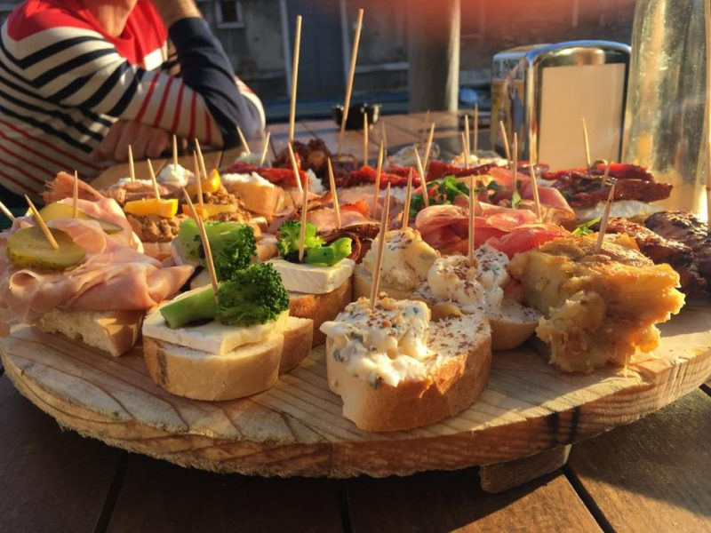 Round wooden platter of colourful cicchetti topped with cheese, ham, vegetables and toothpicks
