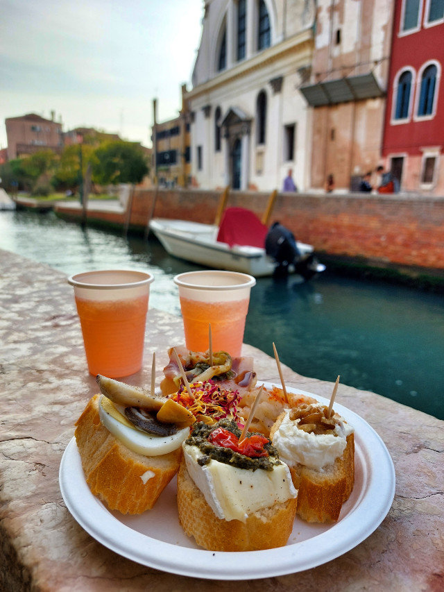 Plate of cicchetti and two orange spritz drinks on a stone ledge beside a canal with boats